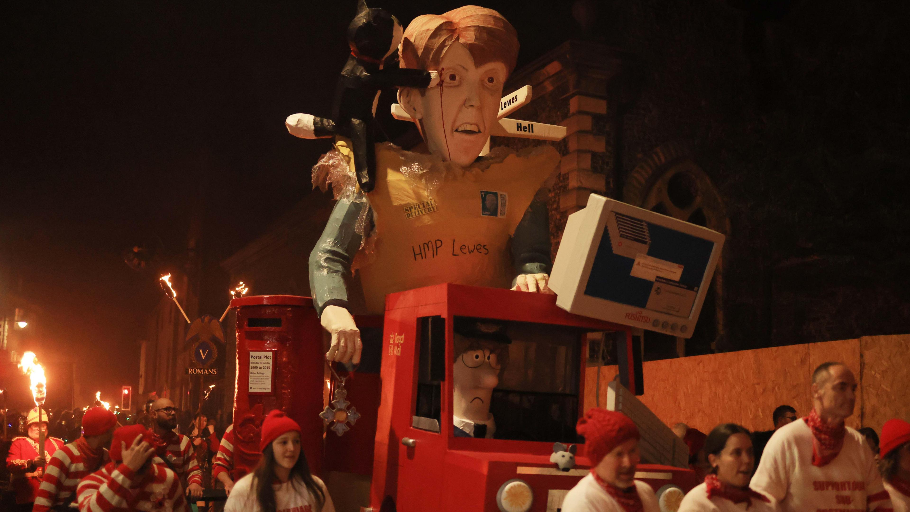 An image of a large effigy of a woman on top of a Post Office van driven by the cartoon character Postman Pat and including a postbox. There are people around the display wearing red and white striped clothing and there are flame torches in the background.