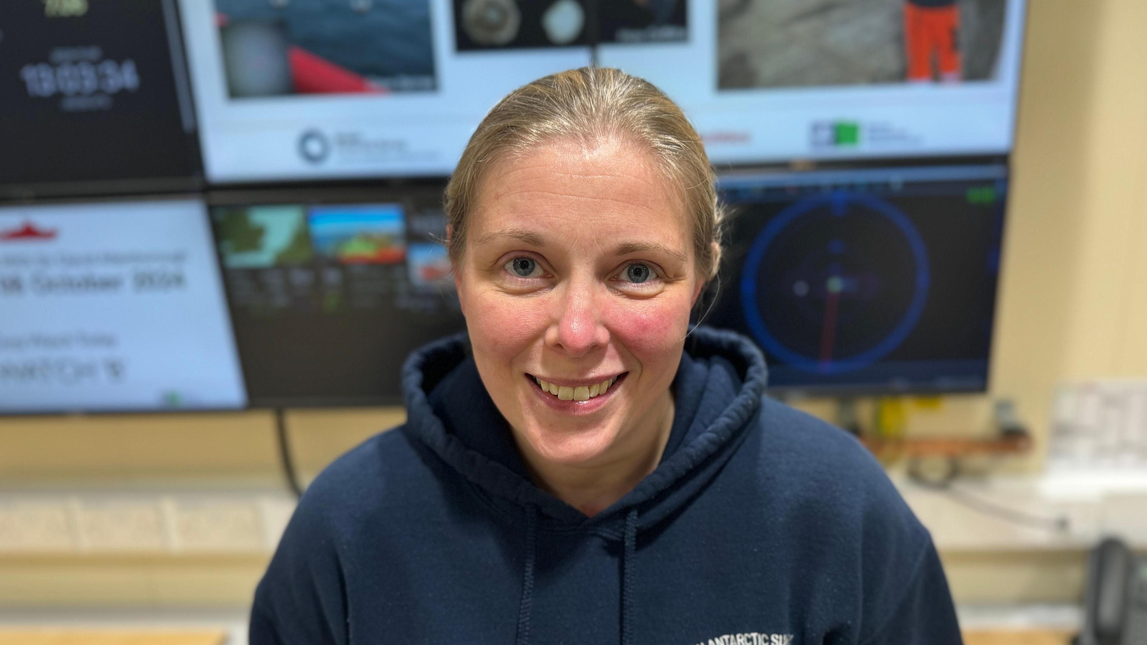 A smiling Natalie Ensor looking straight at the camera. She is wearing a blue hoodie and is standing in front of several computer monitors.