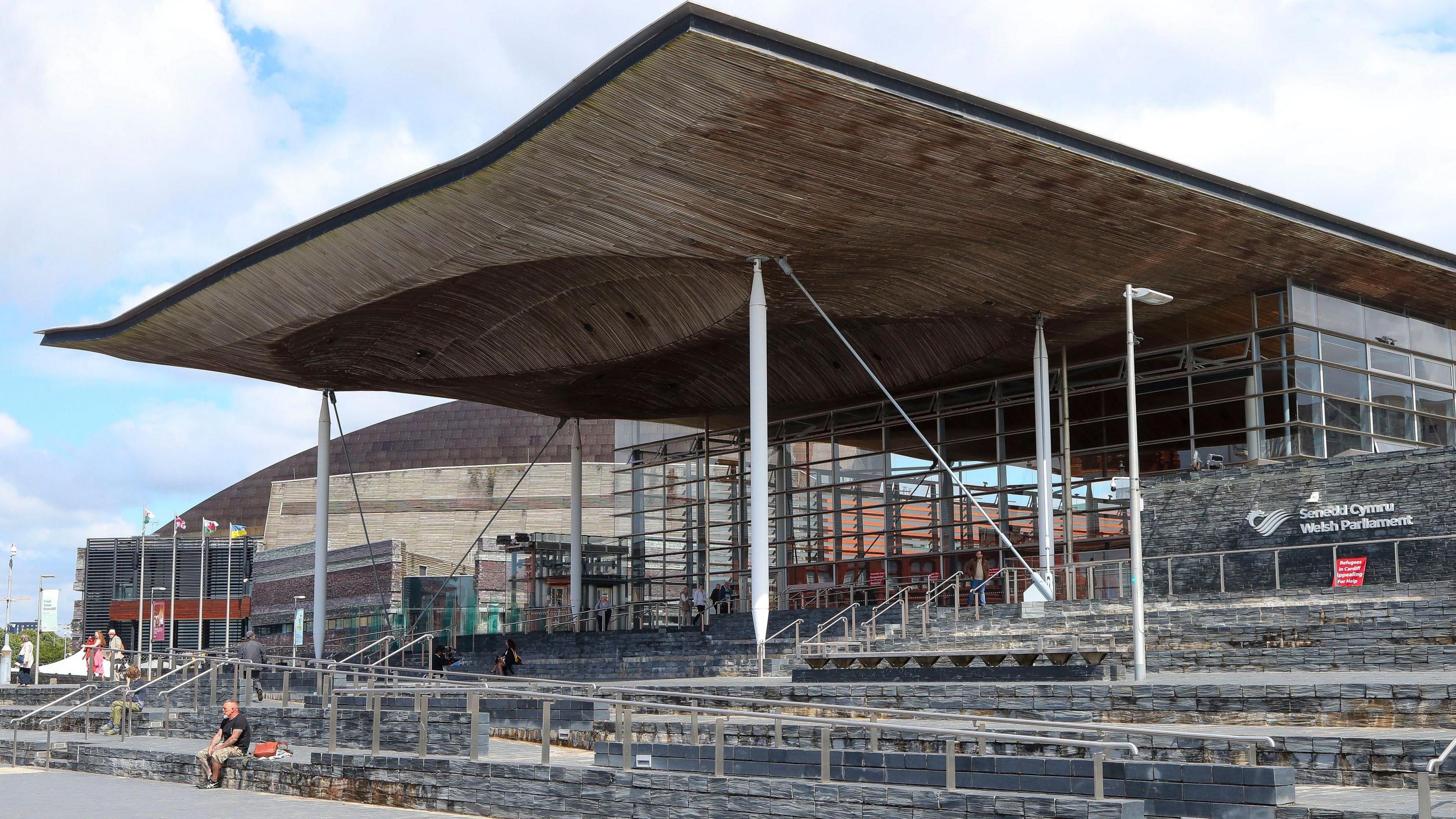 The Senedd building in Cardiff Bay on a bright, warm day. A large roof extends over the steps at the front of the building and the words Senedd Cymru Welsh Parliament can be seen on a wall on the right of the photograph.