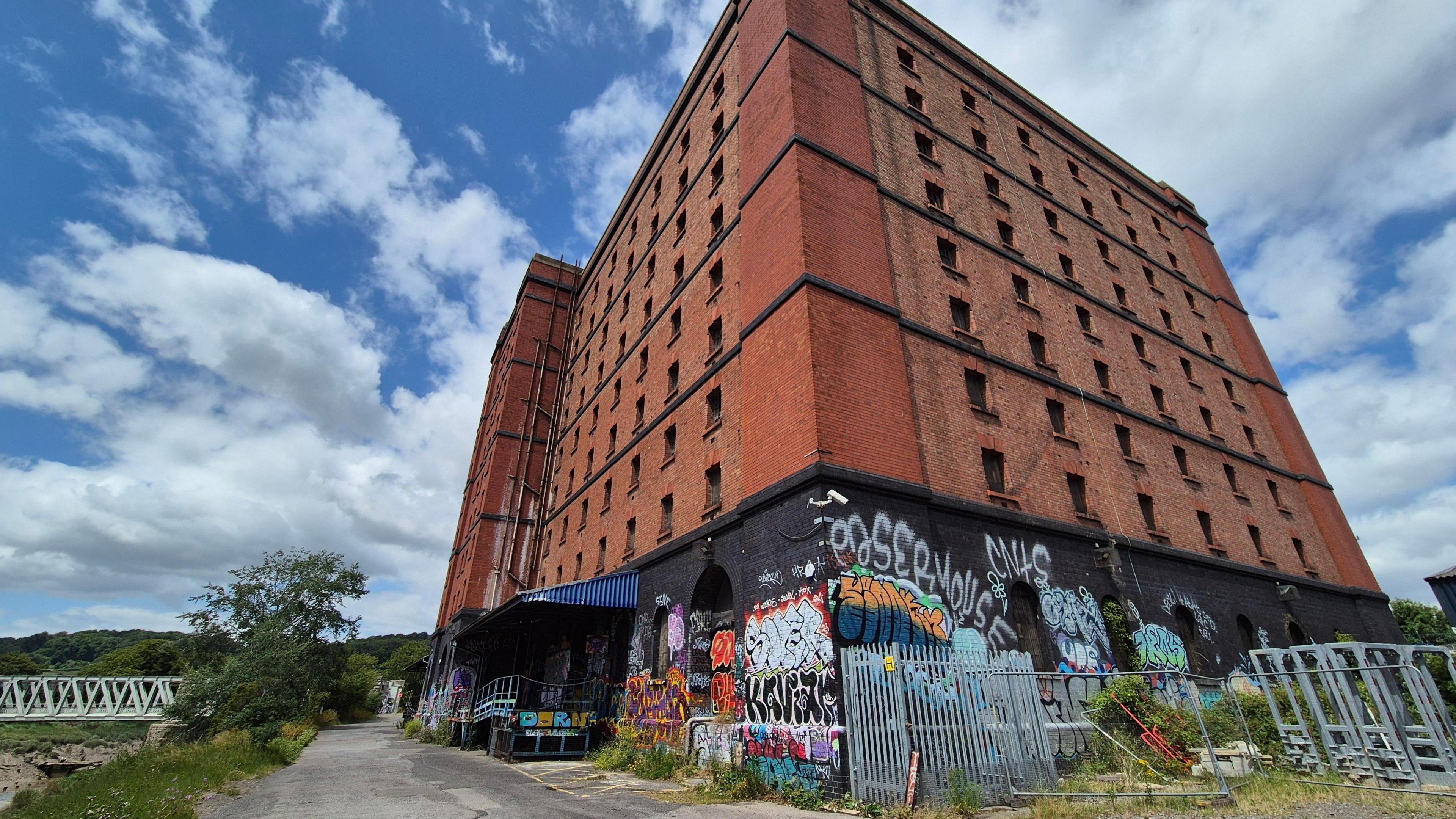 A large red brick warehouse with graffitied walls at the bottom, and a path leading up to a bridge