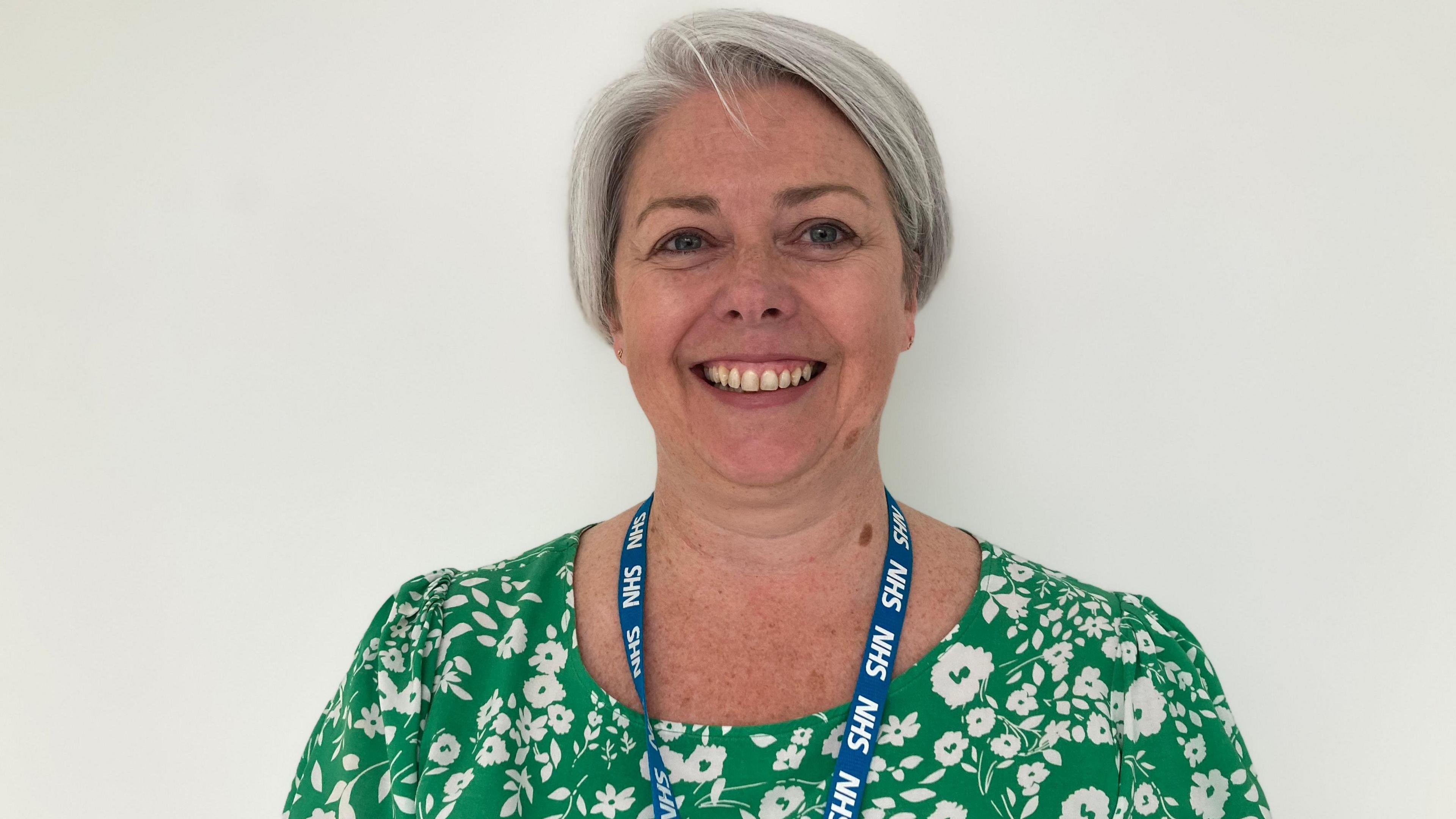 A woman is smiling into the camera. She has short grey hair and is wearing a green and white floral dress and an NHS lanyard is around her neck. She's standing with a white wall behind her.