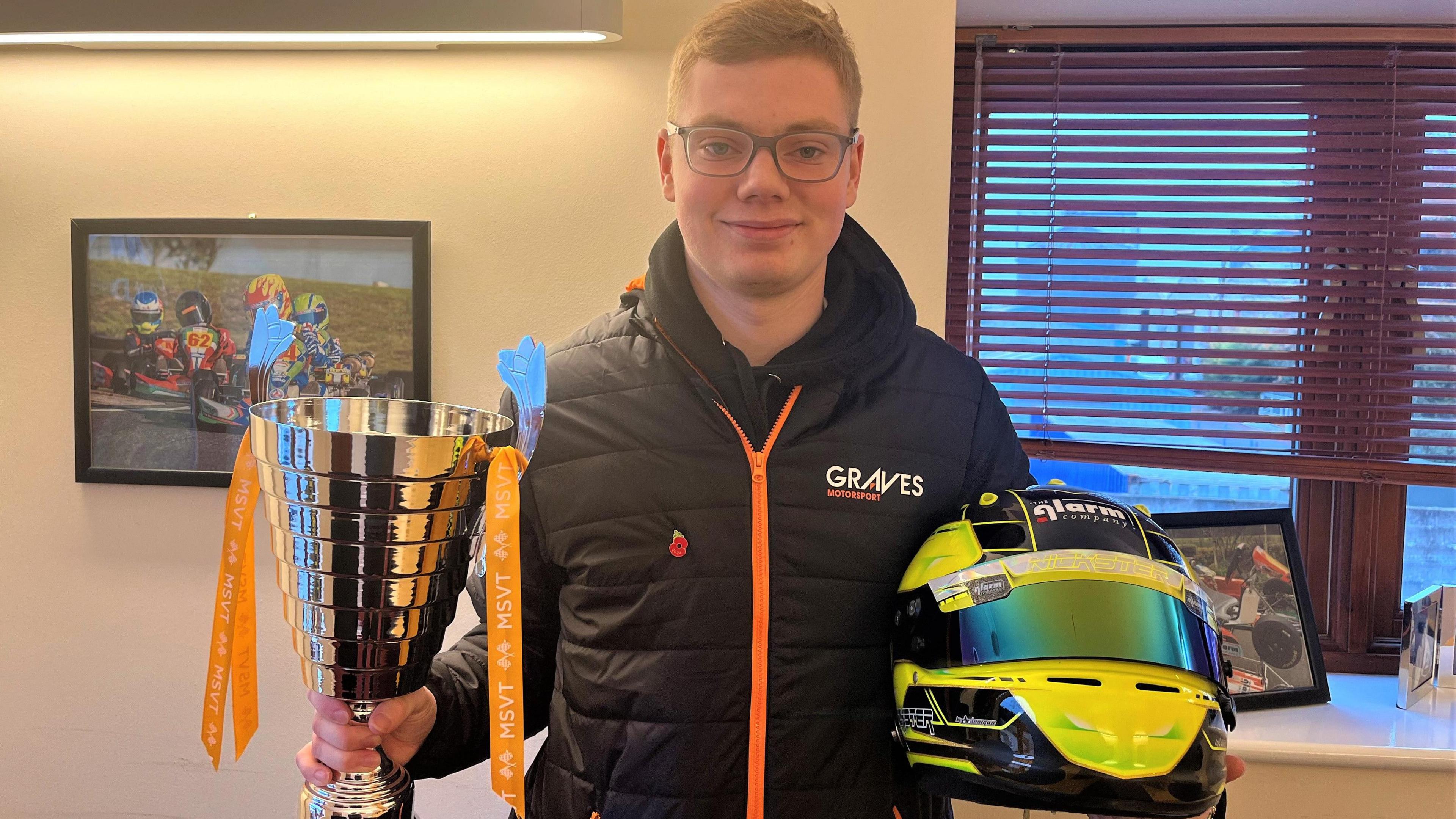 Nicky Taylor stands holding his Clio Cup Trophy and helmet, in a Graves motorsport-branded thermal. Nicky is posing in an office, with a picture of go-karting on the wall behind him.