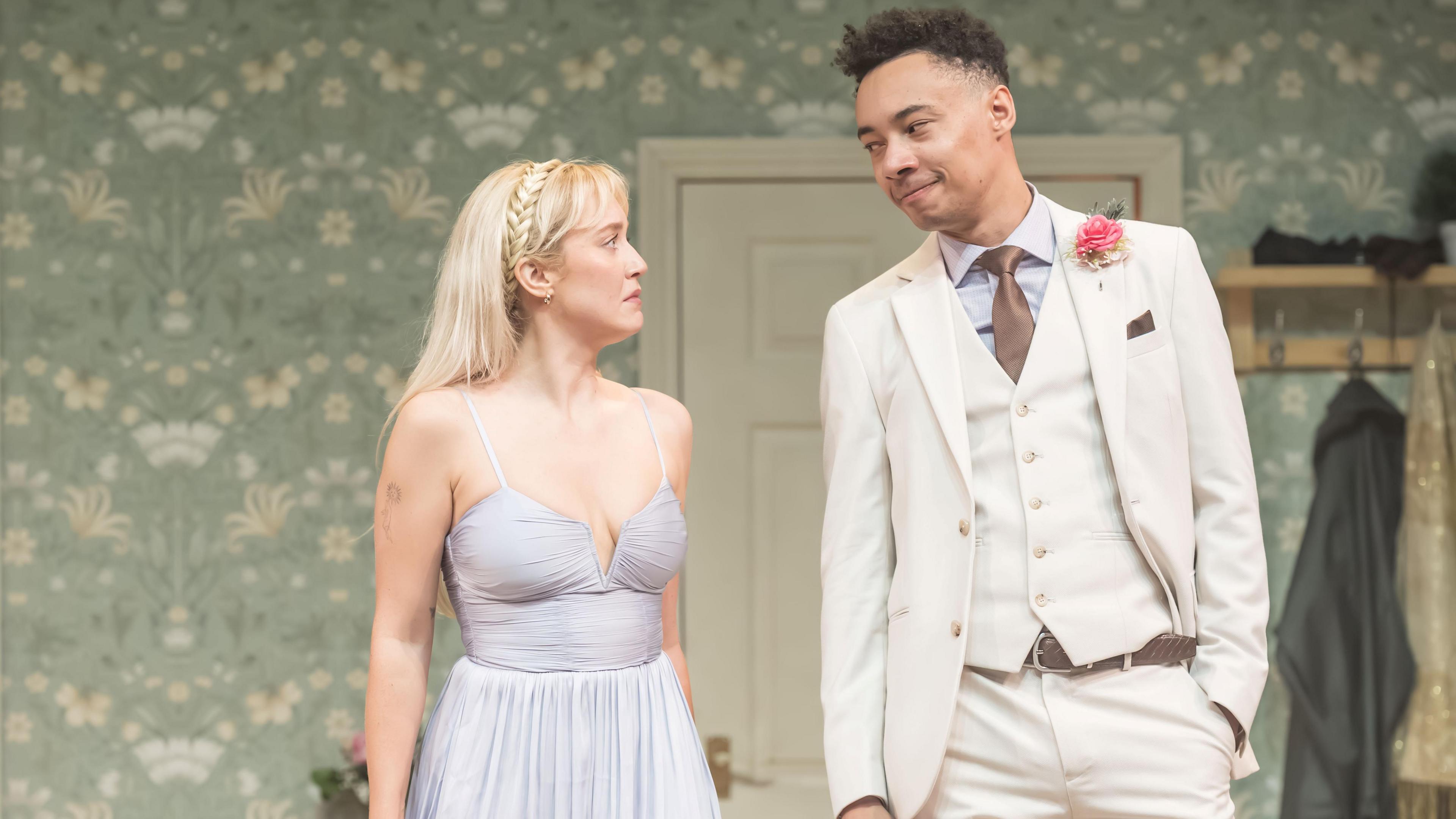 Amelia Kenworthy and Kit Young star in Pickett's Emma. Kenworthy is wearing a light blue formal dress and Young is in a cream suit. They are mid-conversation.