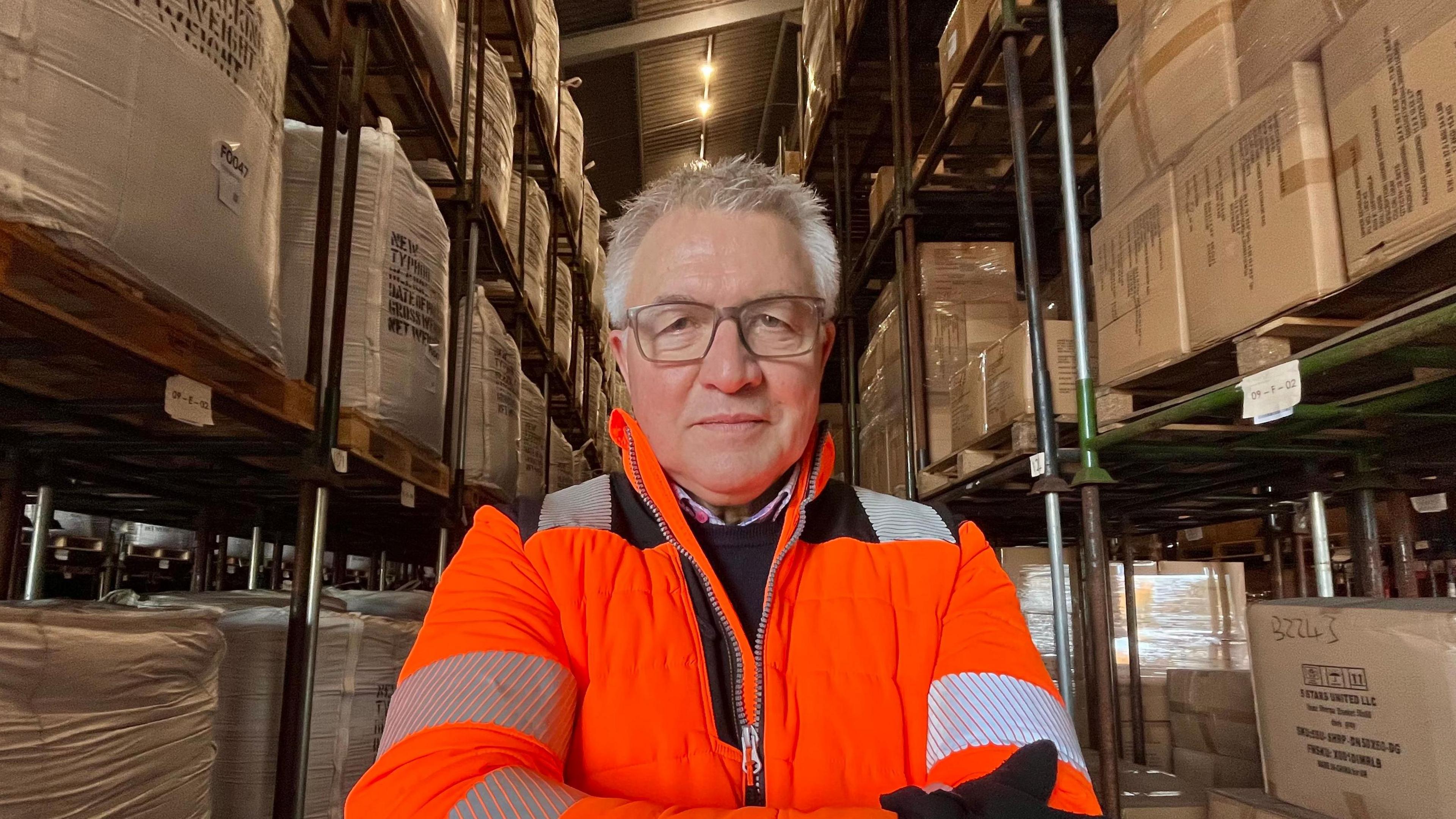 Stephen Britt looks at the camera with an expressionless face. He has short grey hair and wears an orange hi-vis coat. He stands in warehouse with boxes stacked on shelves around him.