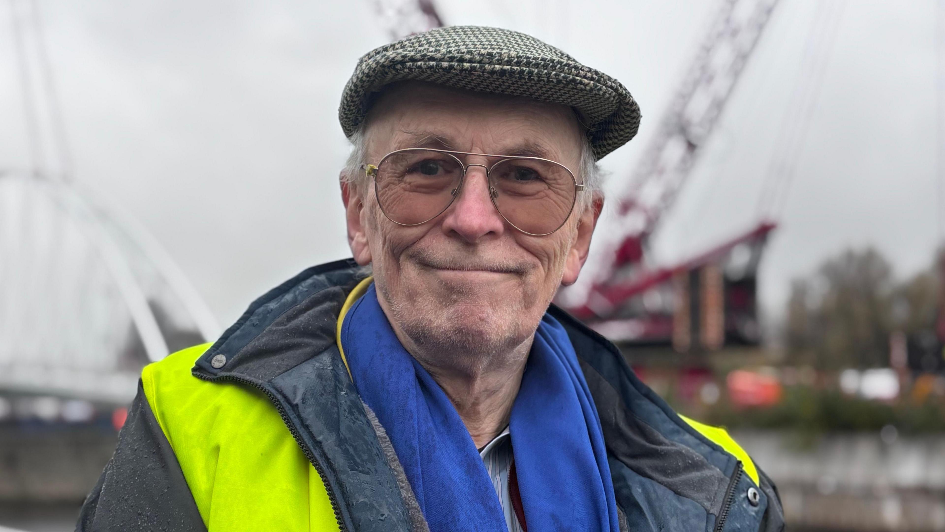 David Nicholson-Cole. An older, White man, wearing a dog-toothed print flat cap and a hi-vis vest over a raincoat. He is also wearing a bright blue scarf. 