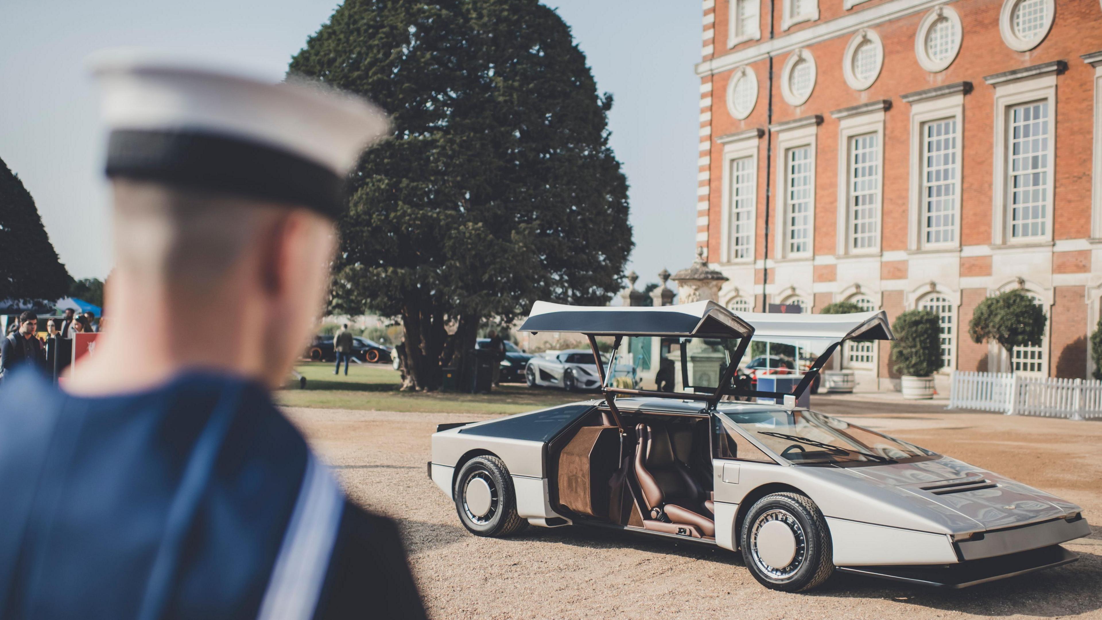 The back of a Royal Naval Air Station apprentice in a blue uniform and hat, he is looking ahead to a cream and beige supercar, it is angular with a pointed nose. the side doors open upwards. it sits parked in front of Hampton Court