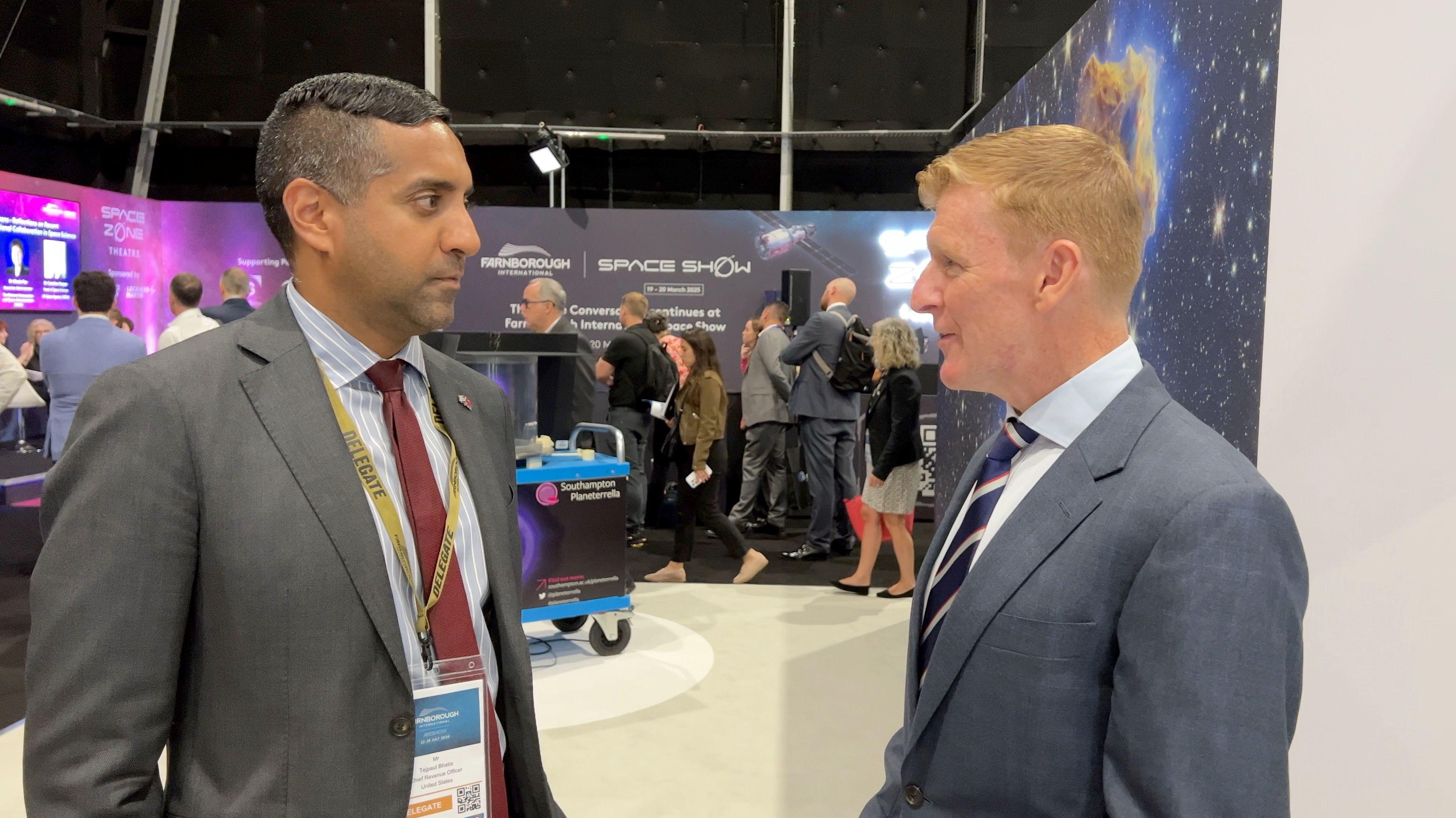 Two men, both in suits, talking to one another with billboards sporting the Farnborough Space Show logo behind them