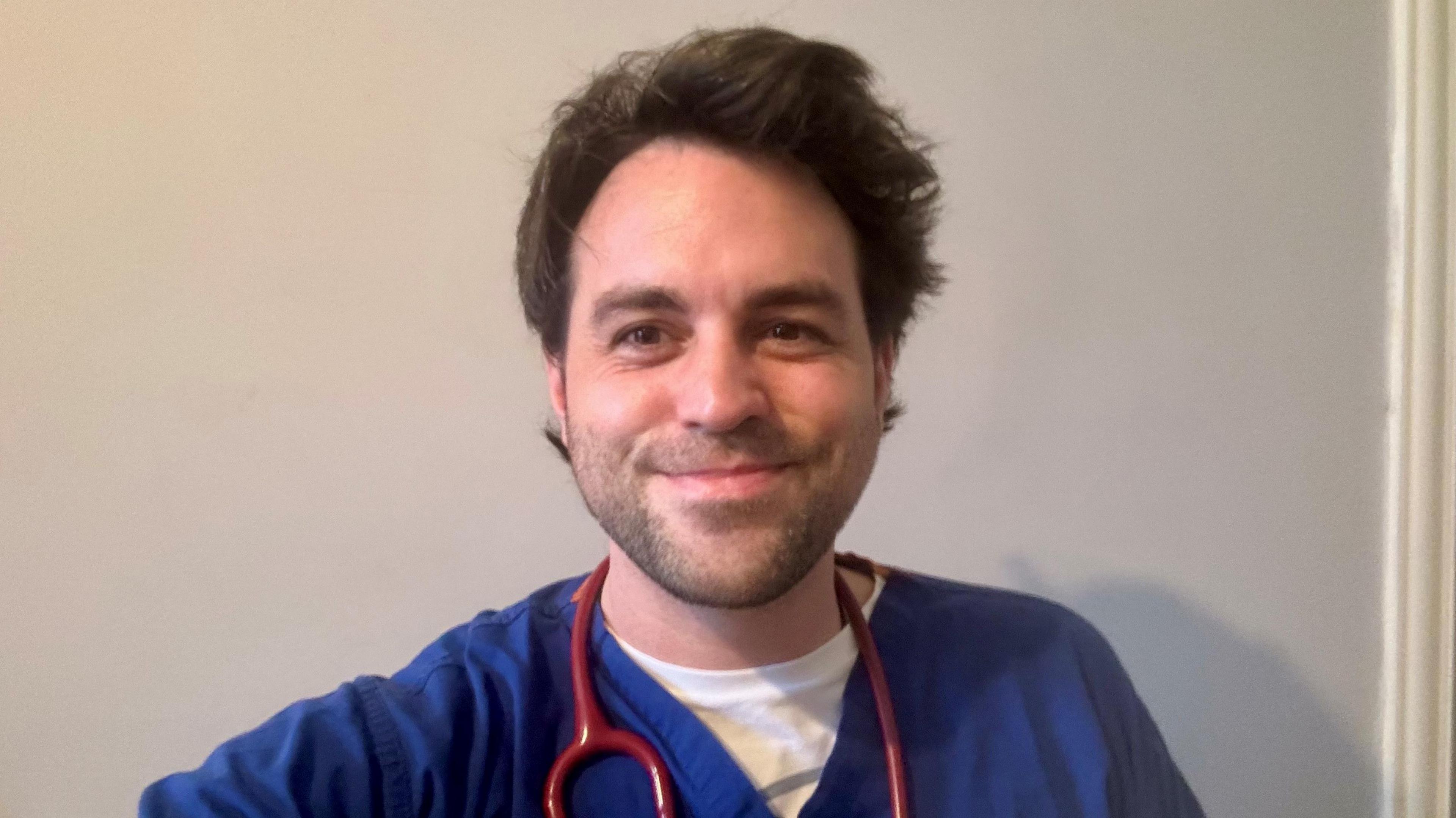 Tommy Collins, a man with dark hair and stubble wears blue scrubs, a white t-shirt and a red stethoscope. 