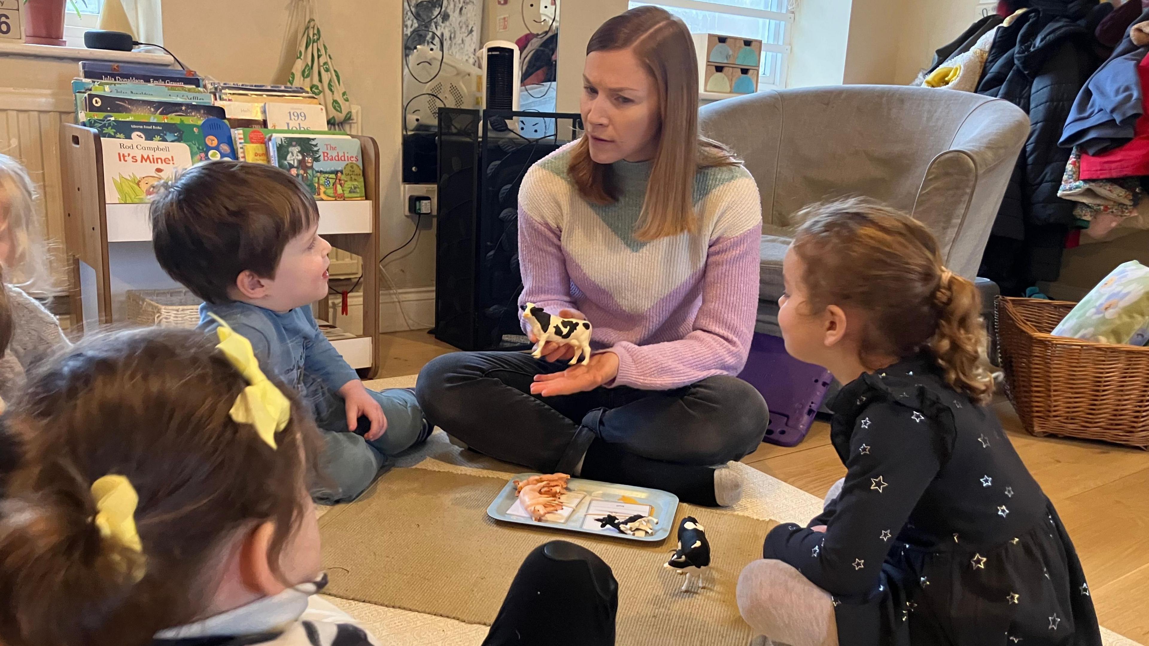 A nursery early years worker shows a group of children sitting in a circle a cow. She has long brown hair and a stripy top on.