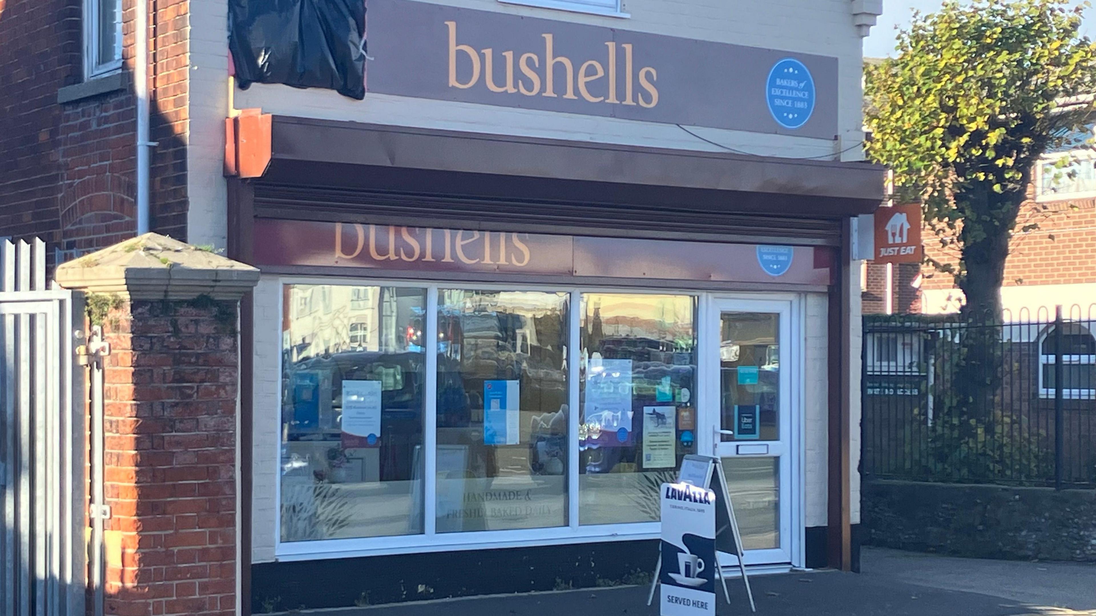 The front of a bakery with purple and white boarding and the name "Bushells" is written in large orange lettering