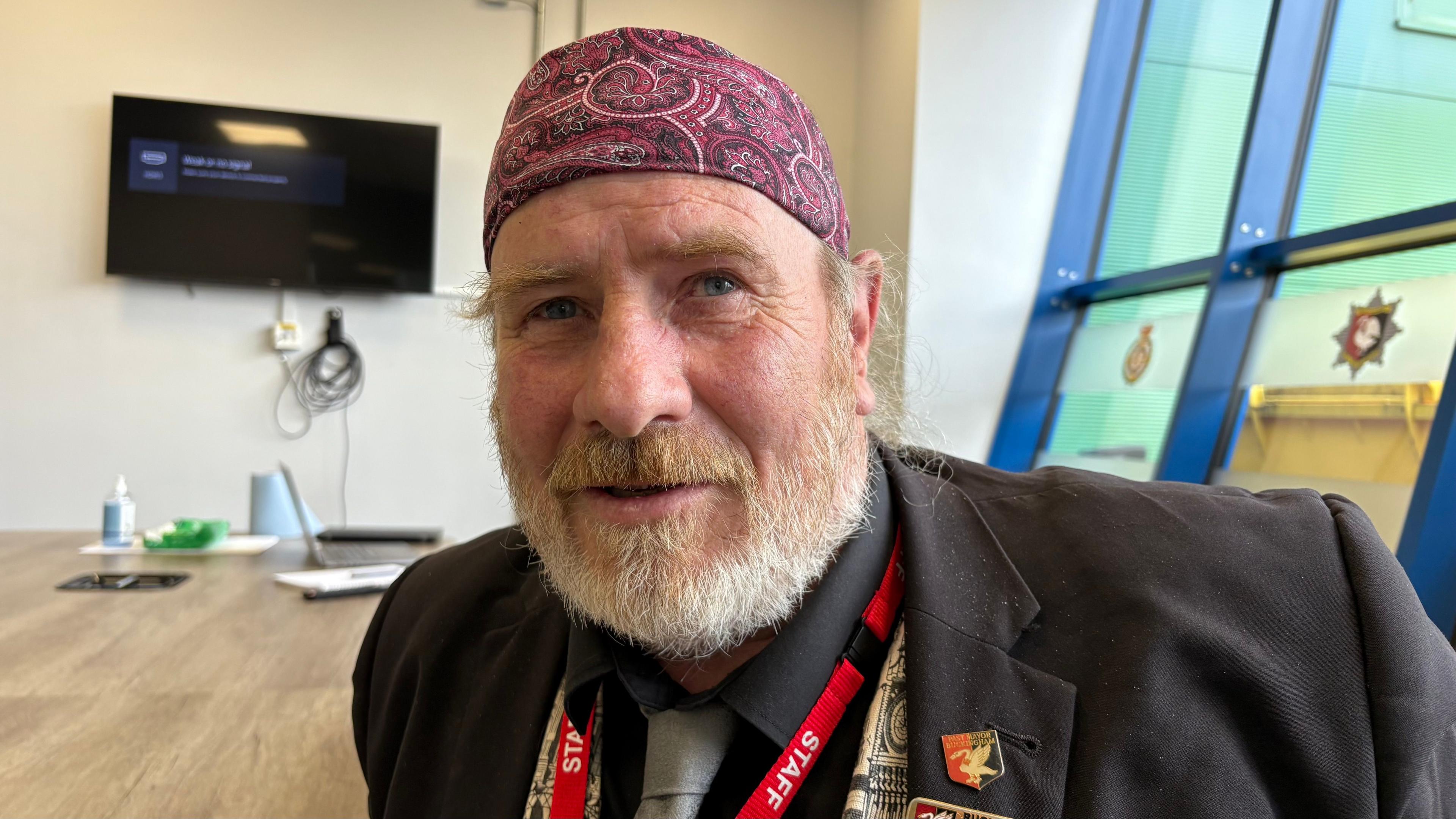 Robin Stutchbury with a ginger and white beard wearing a purple patterned head covering, a dark brown jacket, blue shirt and grey tie. He also has a red STAFF lanyard. He is sitting in a room with a large wooden table, a TV screen on the wall and blue-framed windows behind him.