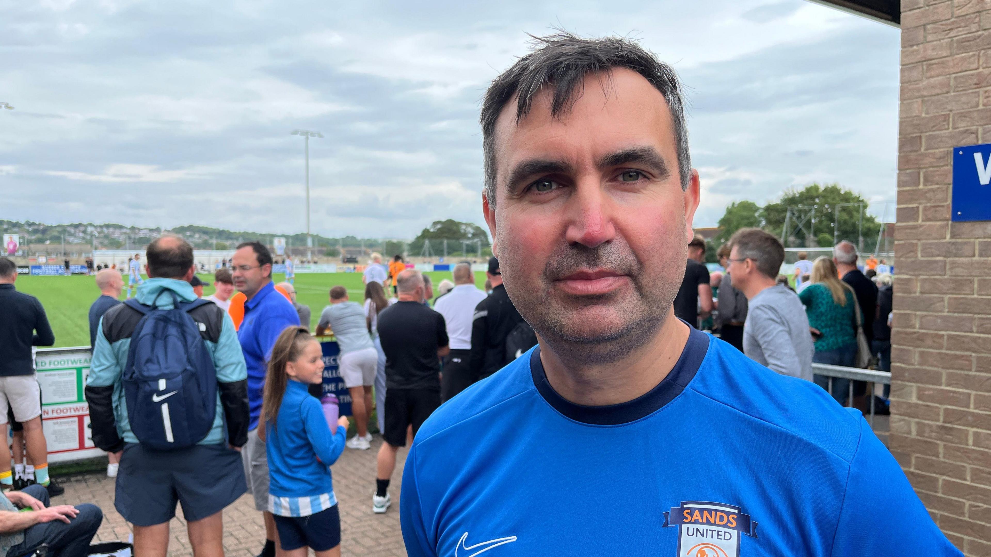 A man wearing a blue football kit stood next to a football pitch. A crowd of people in the background surround the pitch.