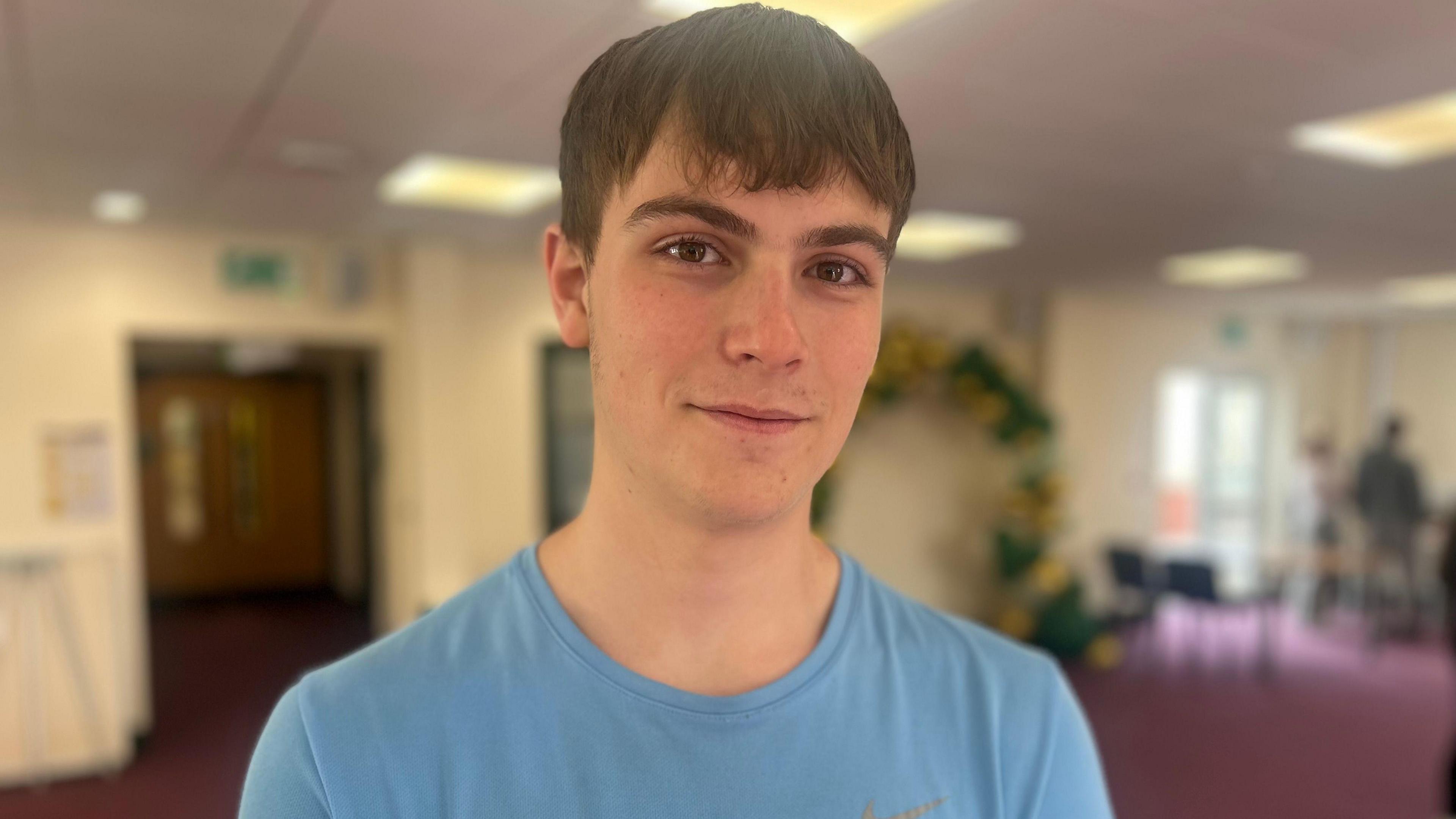 Fynn Stigant wears a blue t-shirt in a classroom. He has short dark hair and smiles.