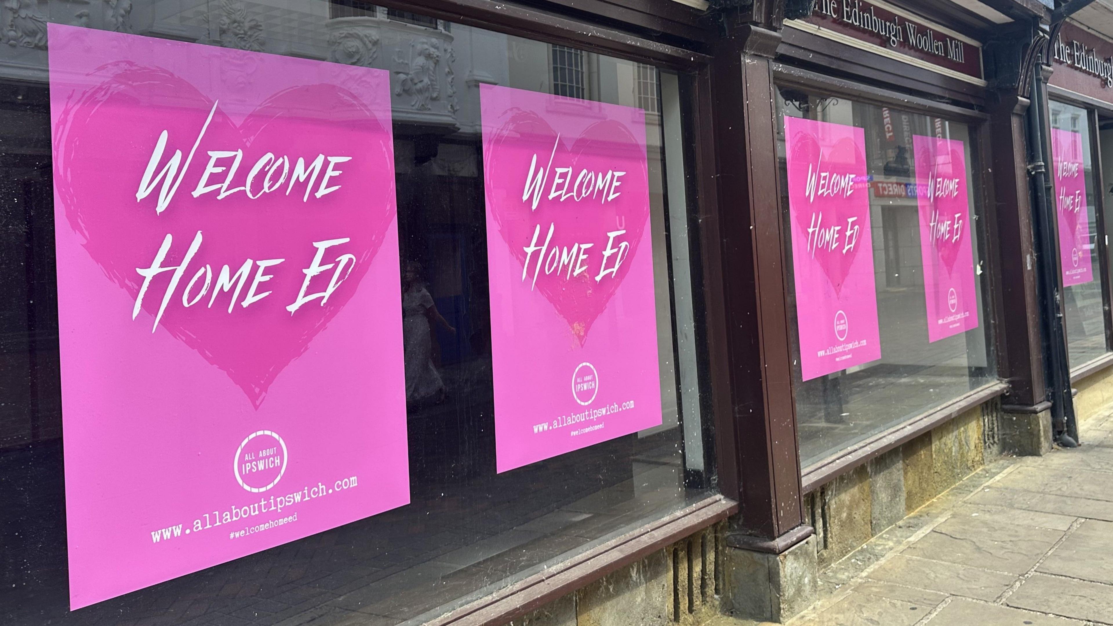 An empty store front with pink posters in the windows that read Welcome Home Ed.