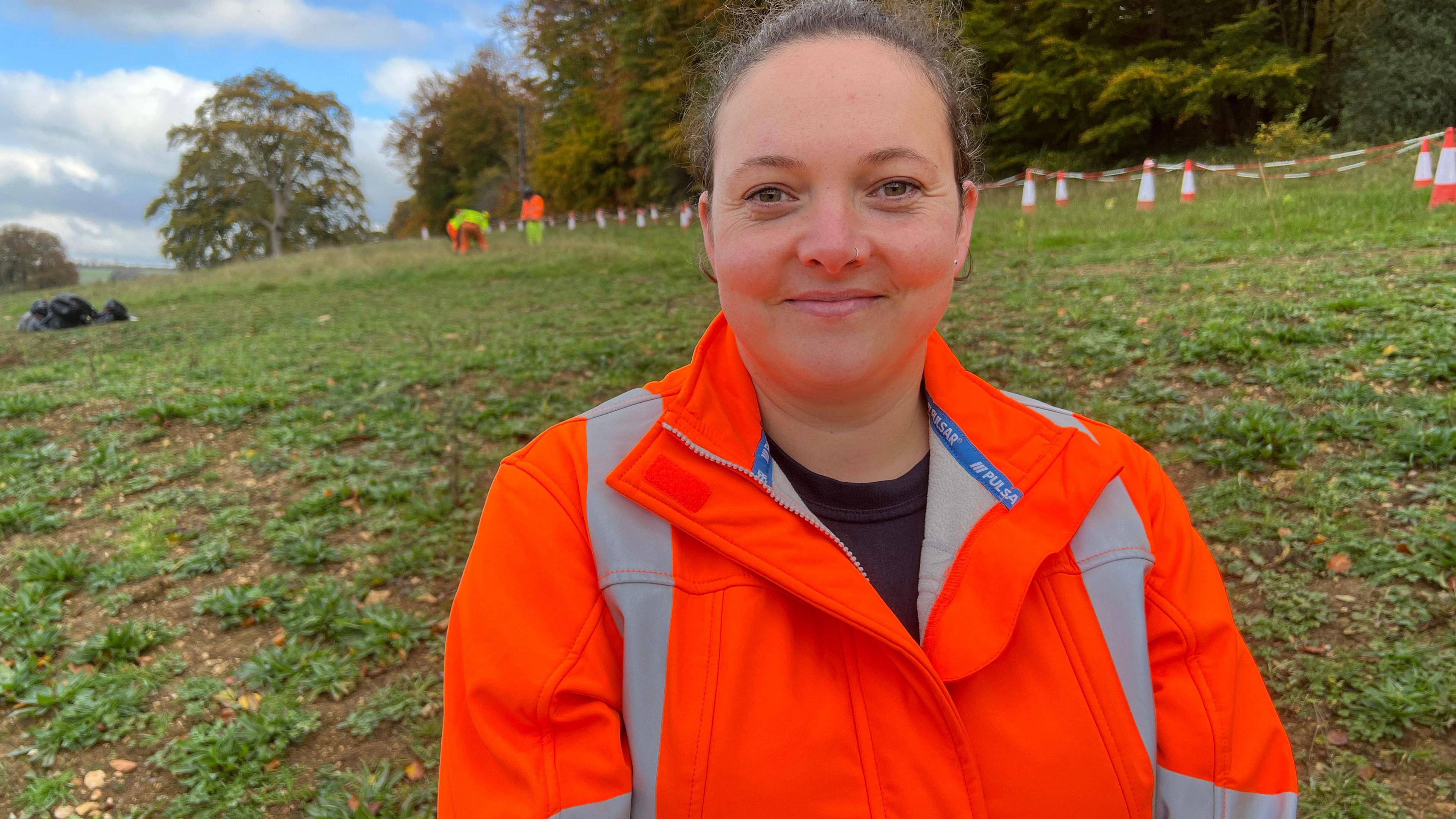 Natasha wearing an orange hi vis jacket smiling at the camera with her hair tied back, standing in front of a team planting trees on a hill