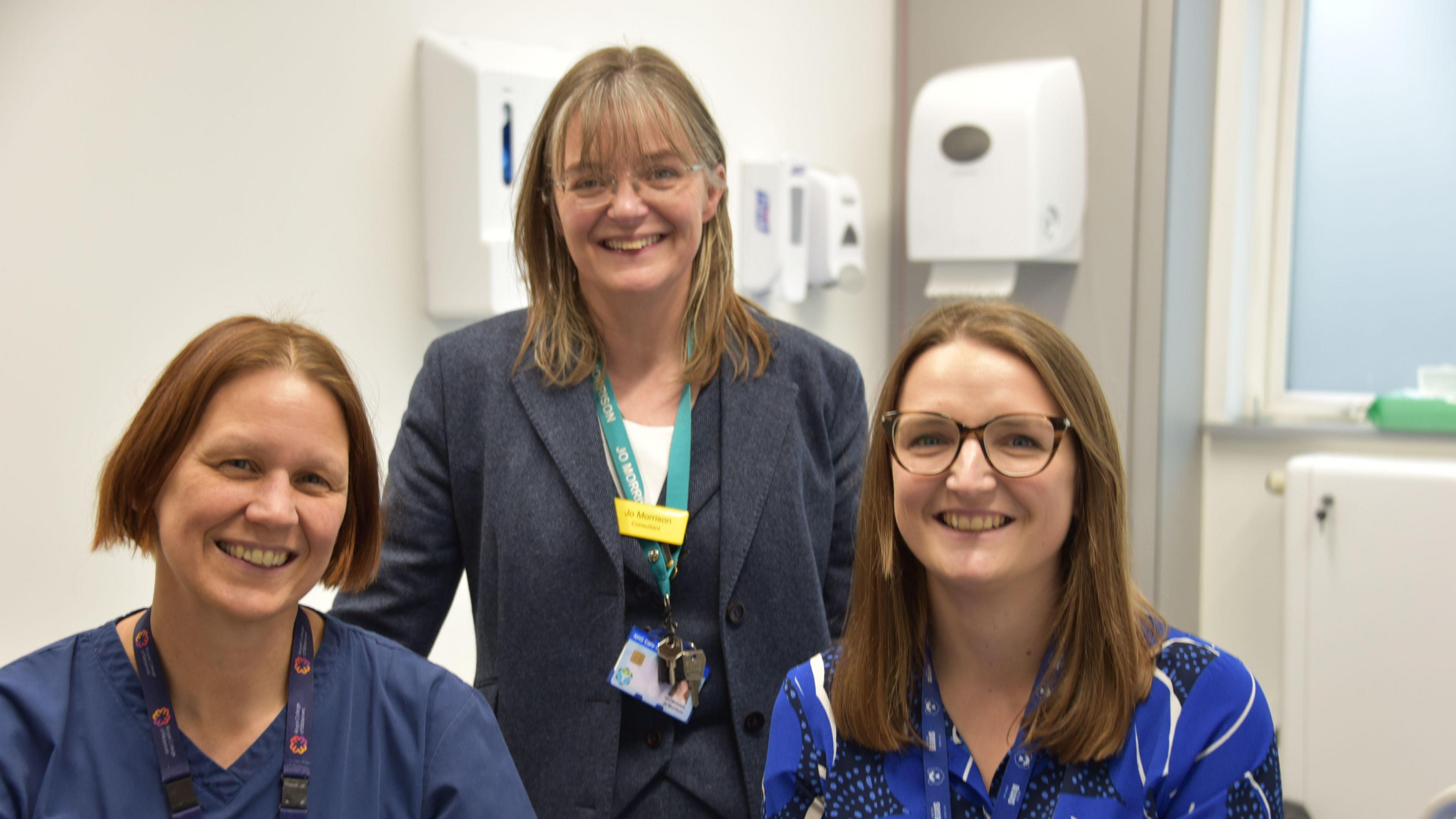 Tessa Dean is on the left of the photo. She has brownish-red hair and is looking at the camera smiling . You can only see her from her armpits up, and is wearing navy blue surgical scrubs. Next to her is Jo Morrison wearing a grey suit. She is stood and pictured from the waist up, with a lanyard around her neck, shoulder length brown hair and glasses on. She is smiling at the camera. Then on the right is Dr Victoria Cullimore. Sheis also smiling at the camera and can be seen from the armpits up. She is wearing a blue blouse, and has brown hair just past her shoulders. They are stood in a white, clinical room. 