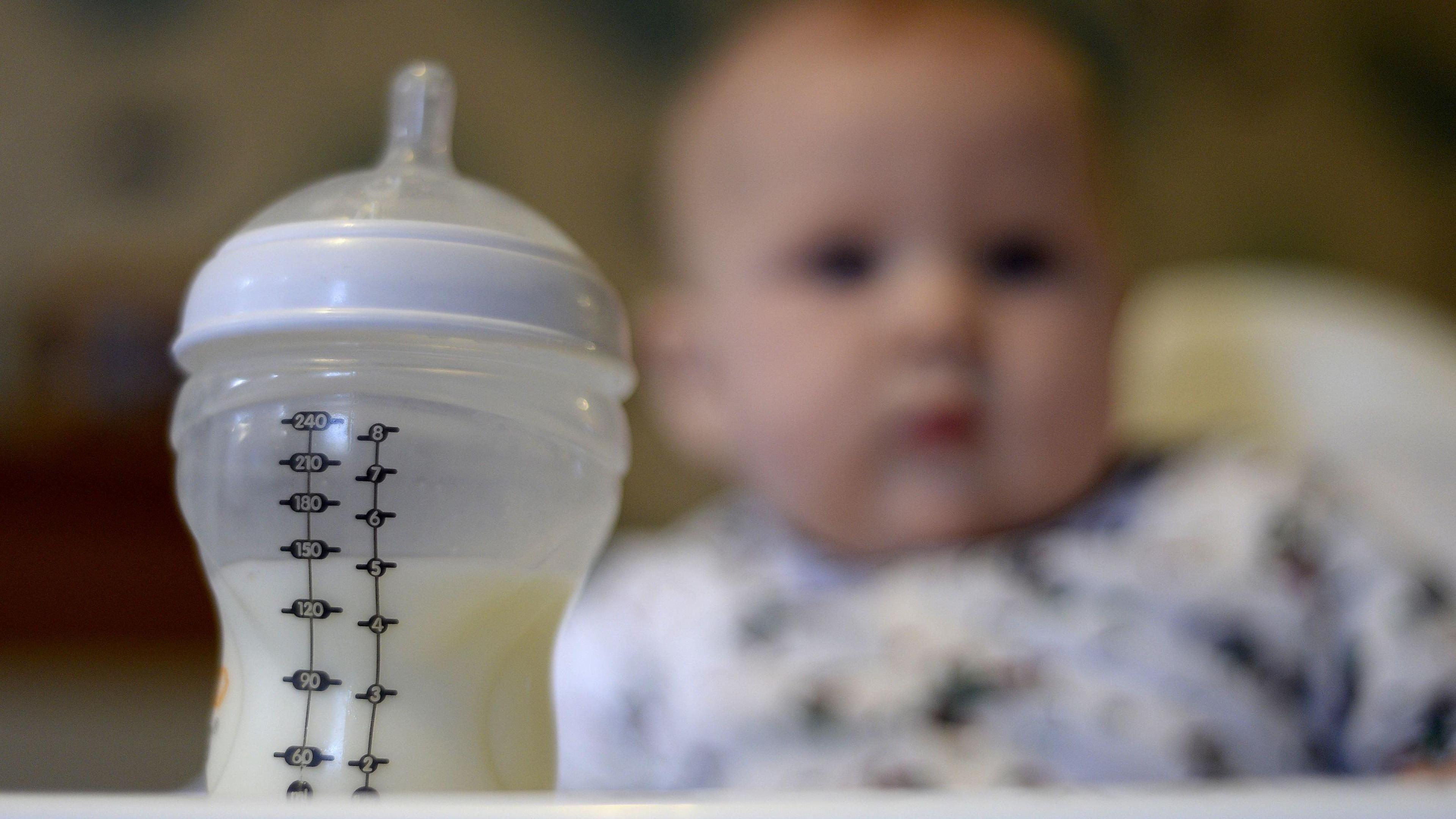 Bottle filled half-filled with baby milk formula with baby sat in a high chair out of focus in the background.