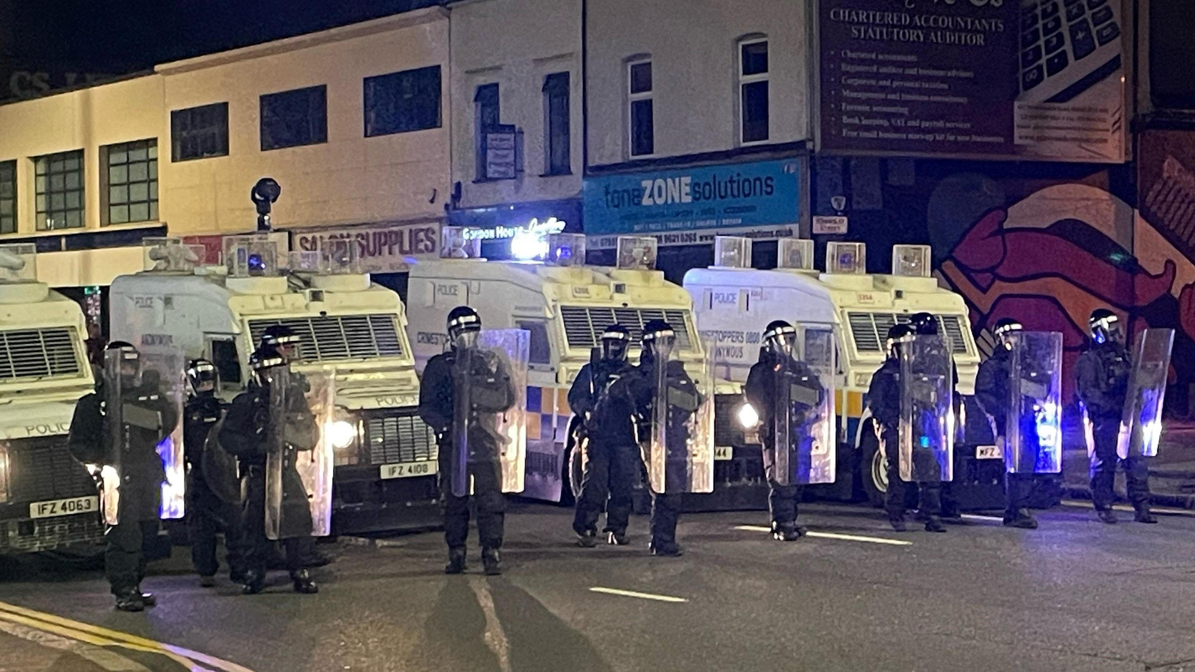 A line of police officers in riot gear, with shields, stand in front of a row of armoured police Land Rovers