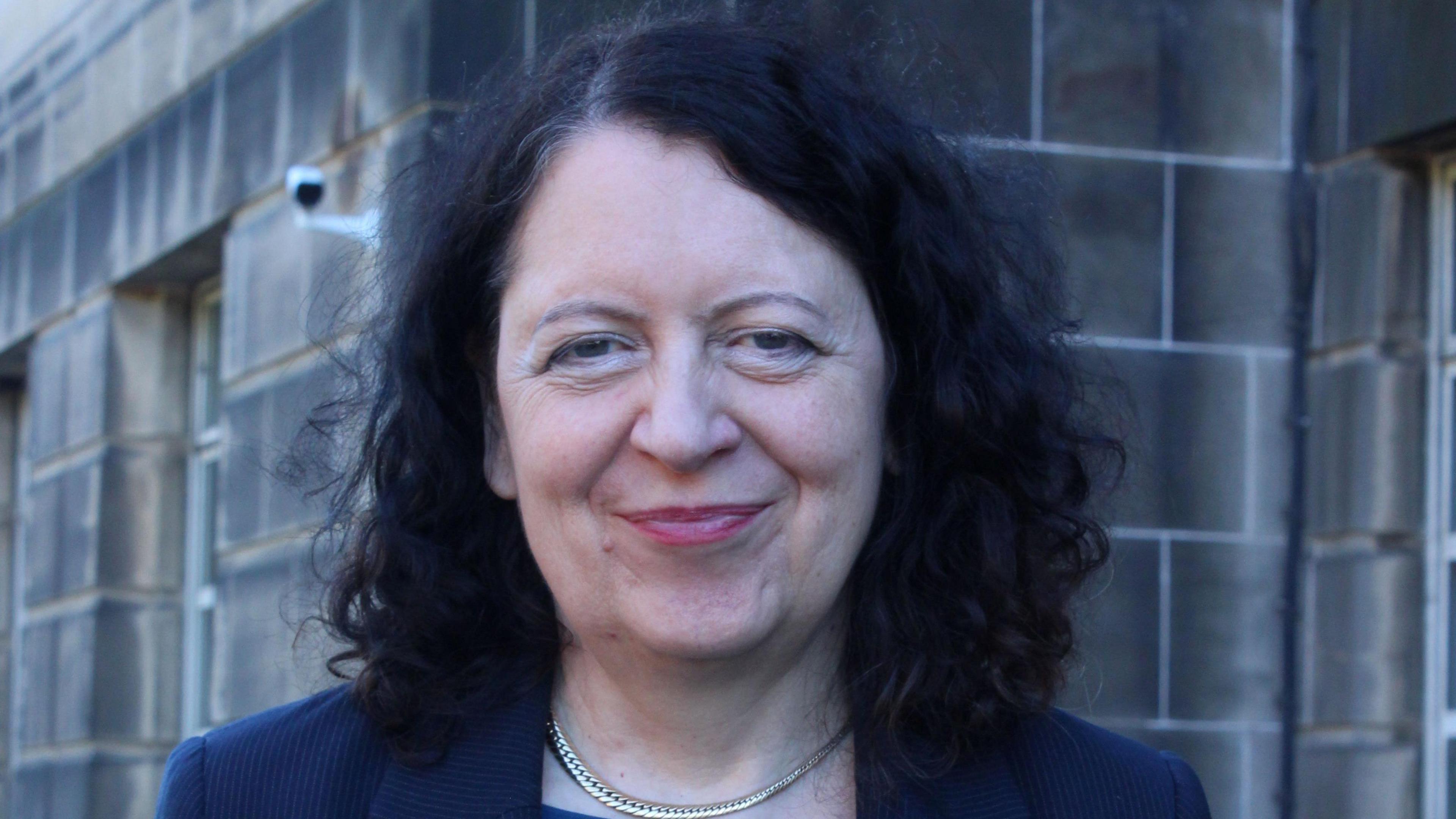 Sheila Duffy is standing in front of a grey sandstone building, looking directly at the camera. She has shoulder length curly black hair and is wearing a blue jacket.