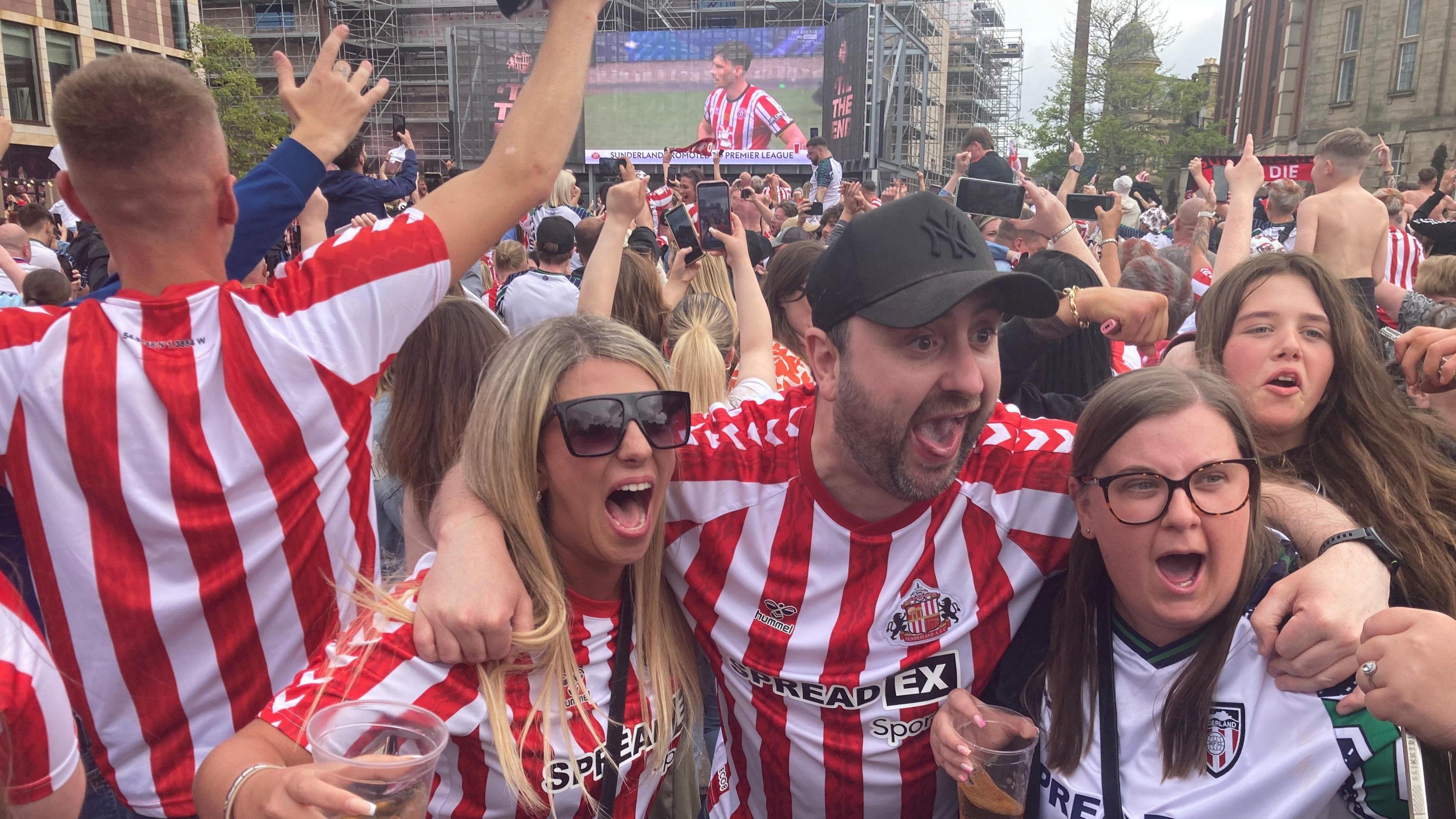 Sunderland fans at the fan zone in the city. Two women and a man - wearing Sunderland colours - are shouting in celebration. Behind them are the backs of fans facing a giant screen. Most have hands raised and many are holding mobile phones aloft.