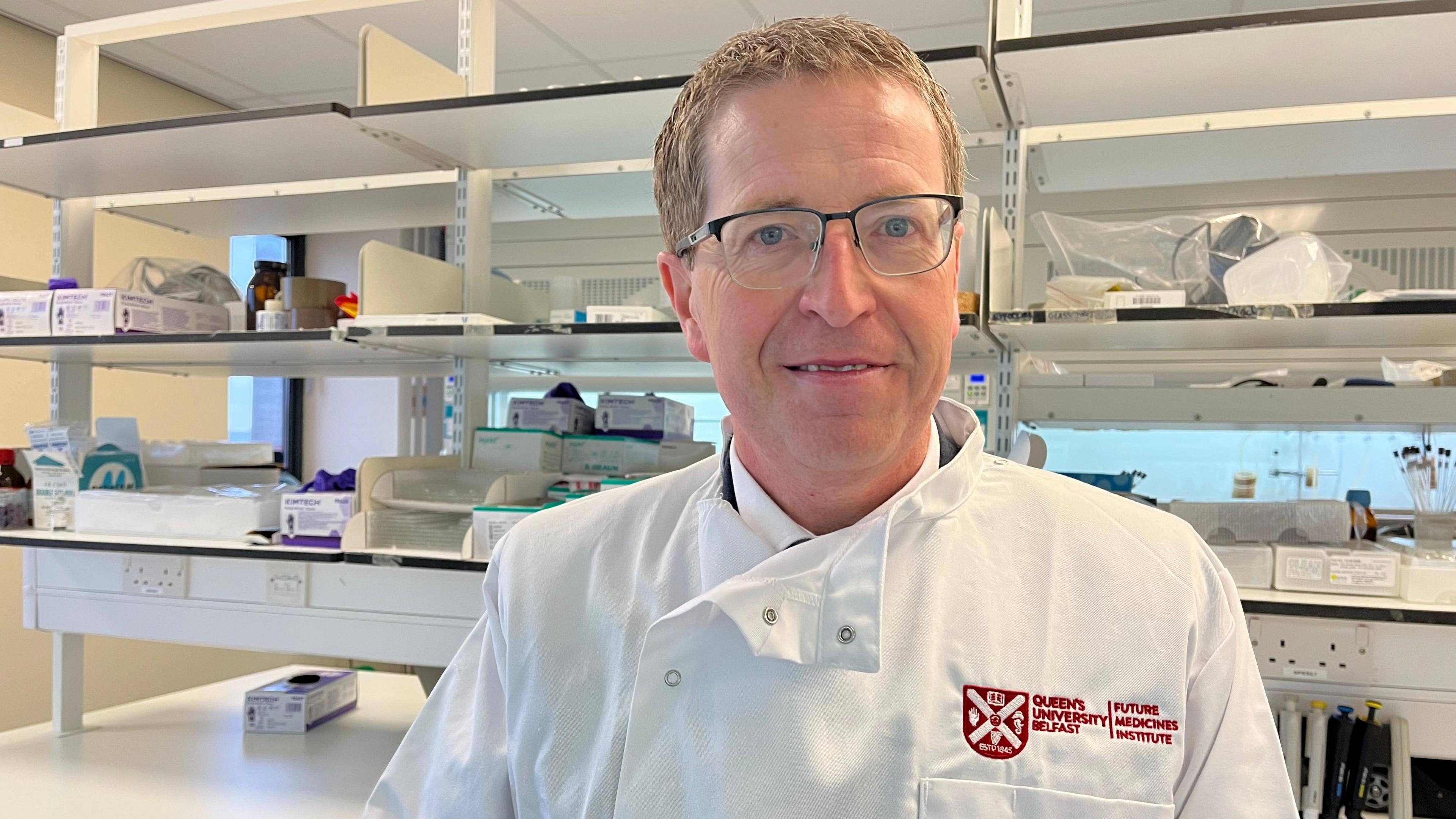 Prof Christopher Scott. He has glasses and short grey-brown hair. He is wearing a white lab coat with a Queen's University Belfast logo on the left breast. In the background there are shelves with various bit of laboratory equipment on them