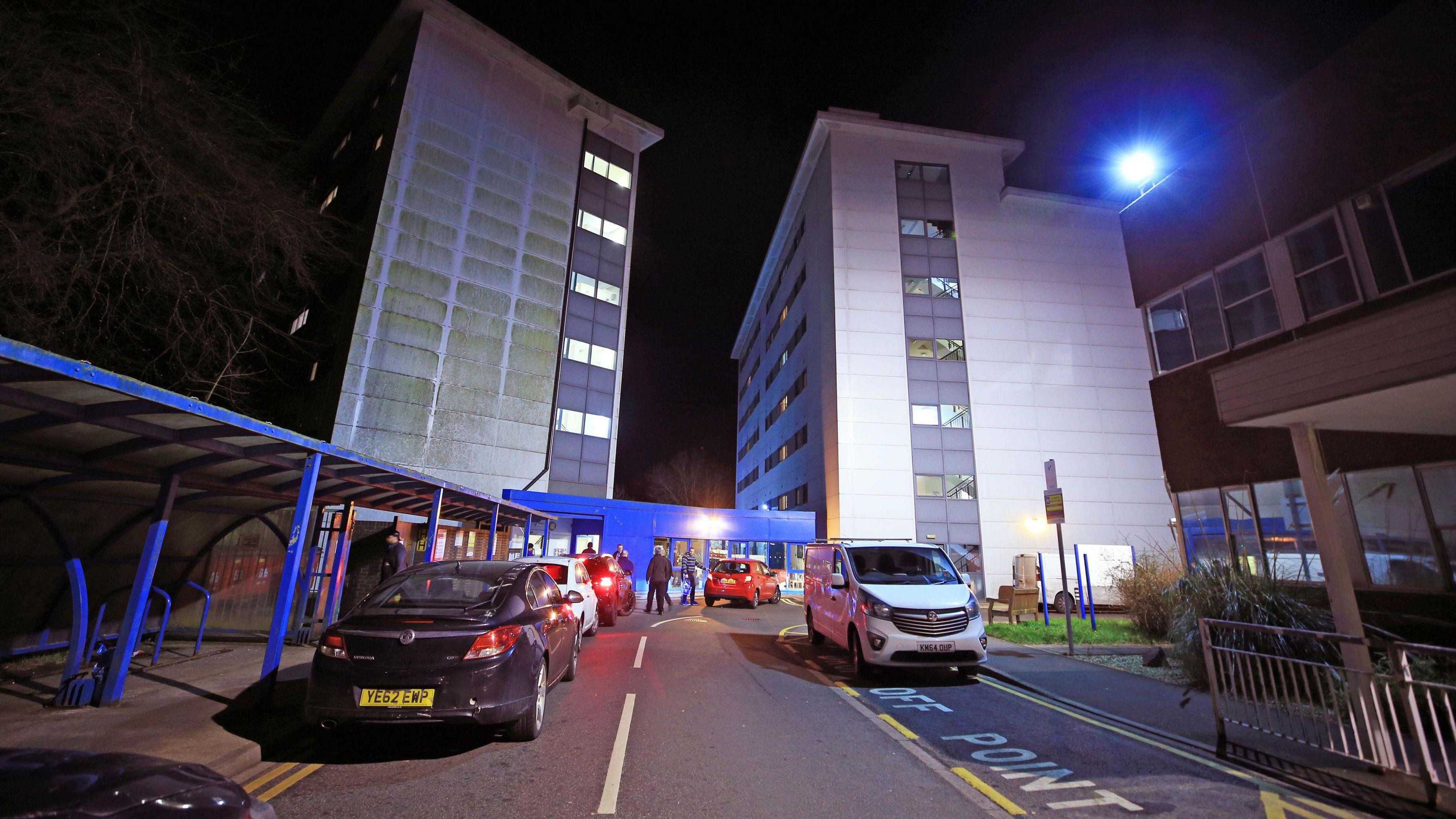 Two, tall grey and white building towers, face each other, with a stretch of road with hospital drop off parking in front of it. It is night time 