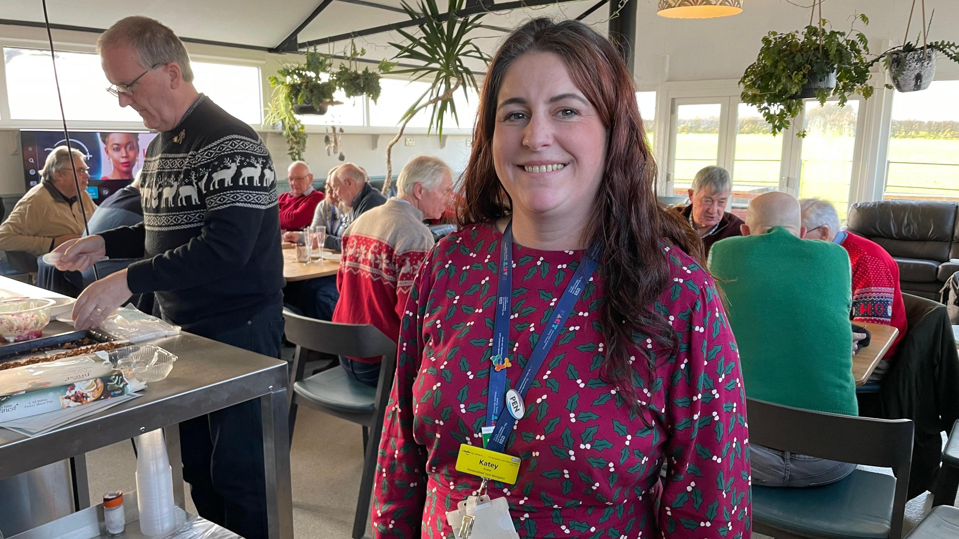 Katey Evans has long red/brown hair and is wearing a maroon red top with green holly leaves on it. She is wearing a dark blue lanyard. In the background are men in Christmas jumpers, sitting together around tables.