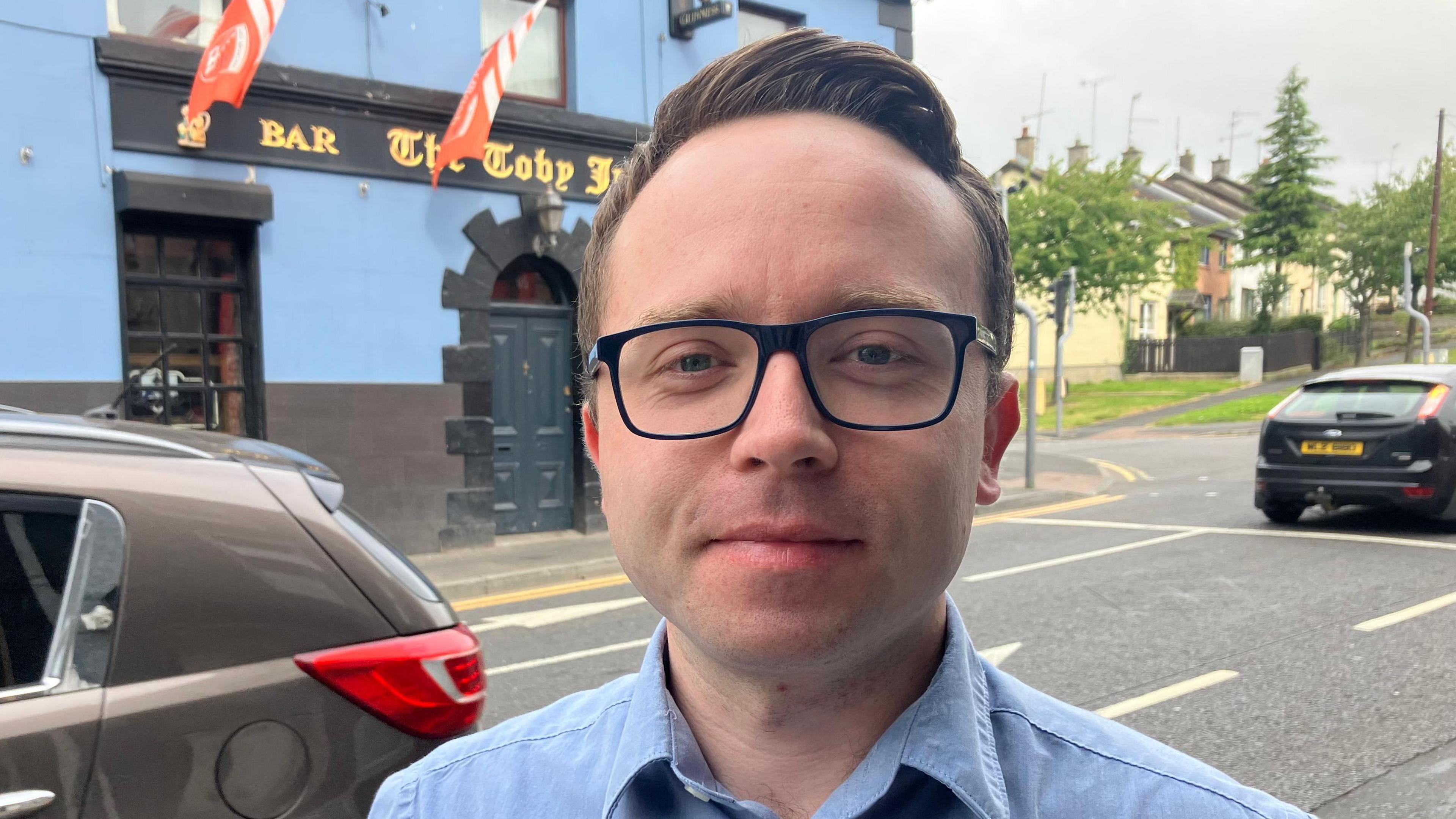 Dáire Hughes has short brown hair, black square glasses and is standing on a street wearing a blue shirt with the top button out. There are cars driving past, one black and one brown.