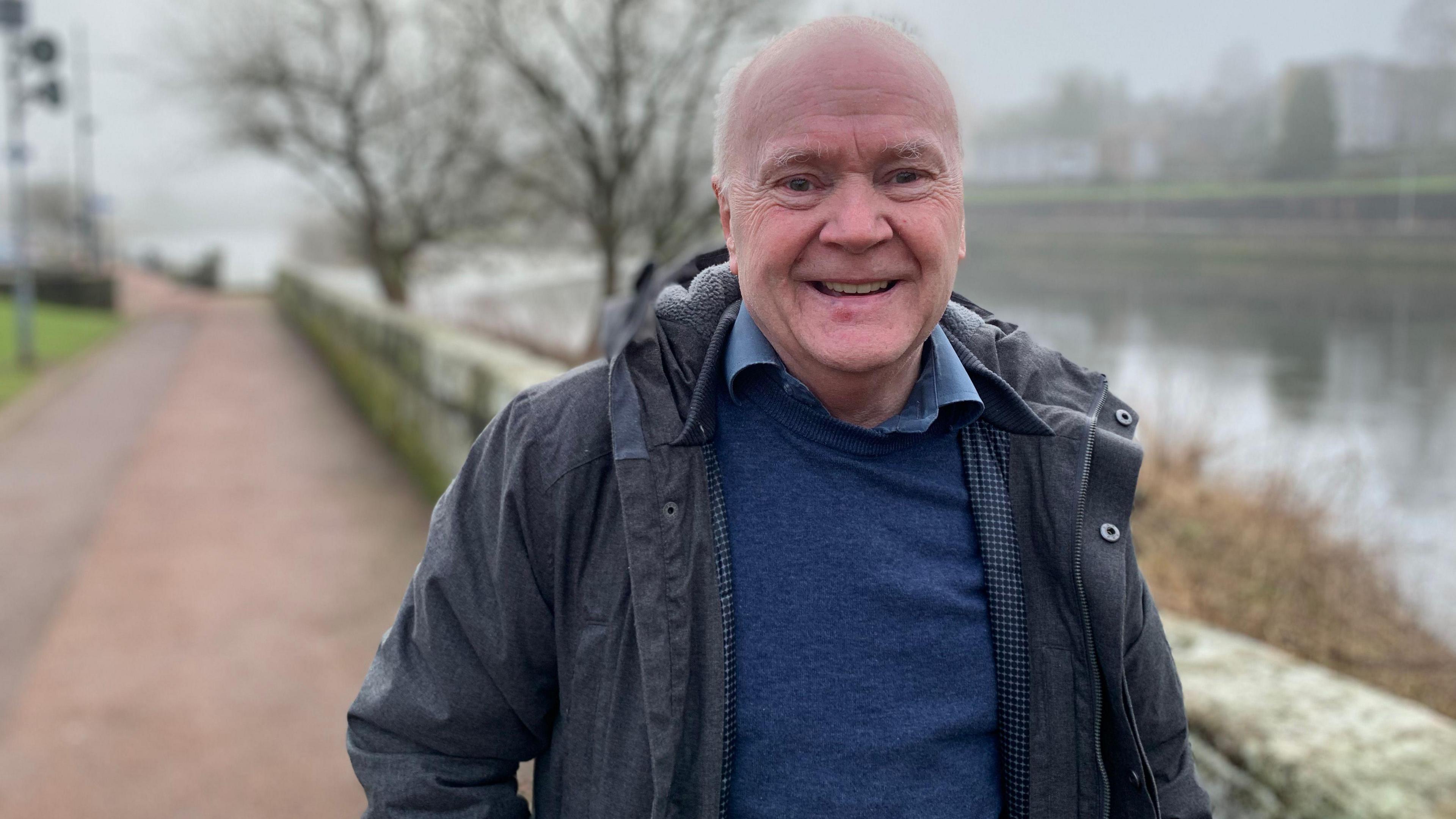 A grey haired, balding man with a dark jacket and blue jumper and shirt stands in front of the River Nith in Dumfries