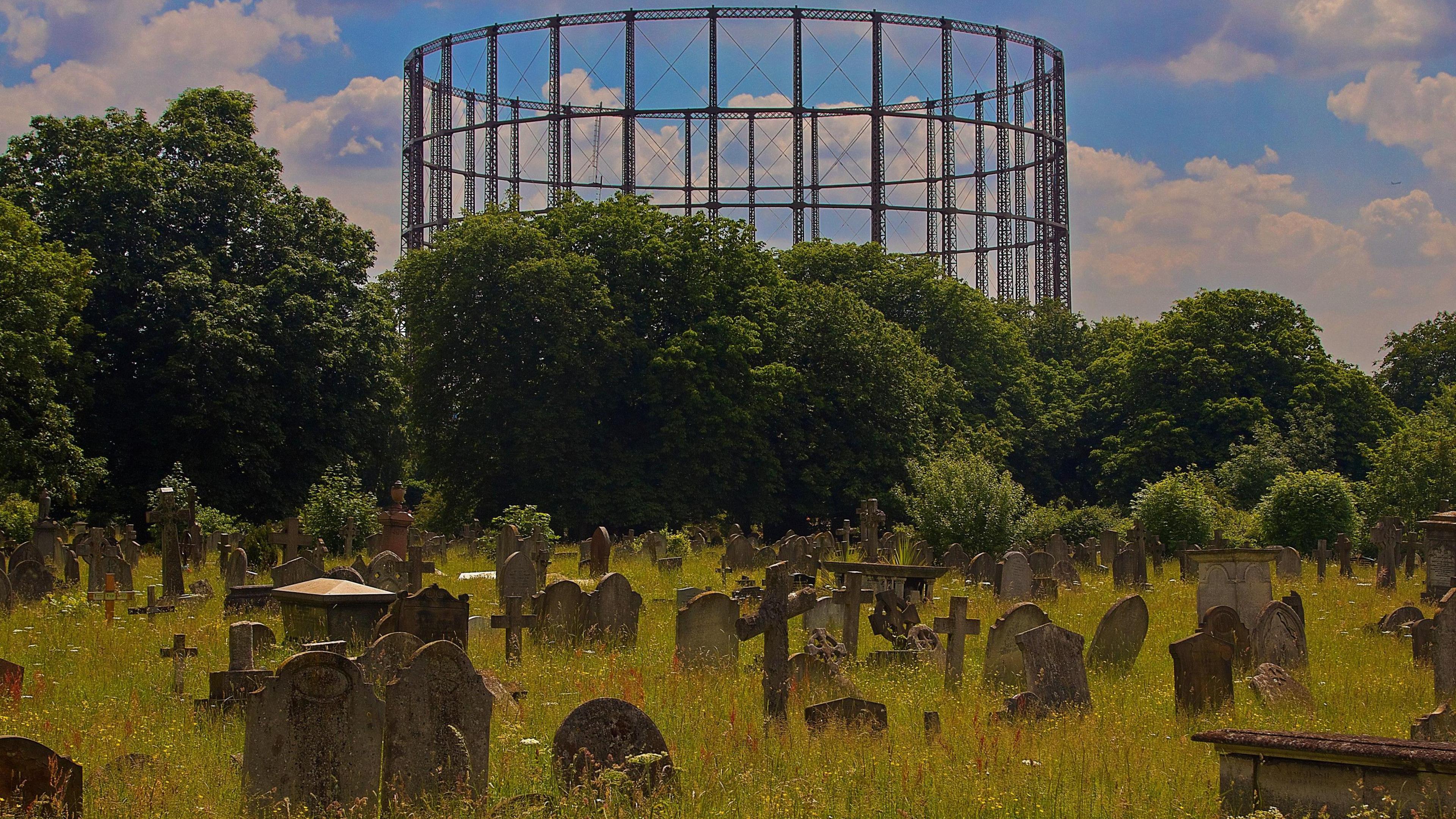 Kensal Green Cemetery with graves hidden in overgrown grass and flowers. In the background are trees with a large gasholder in the skyline.