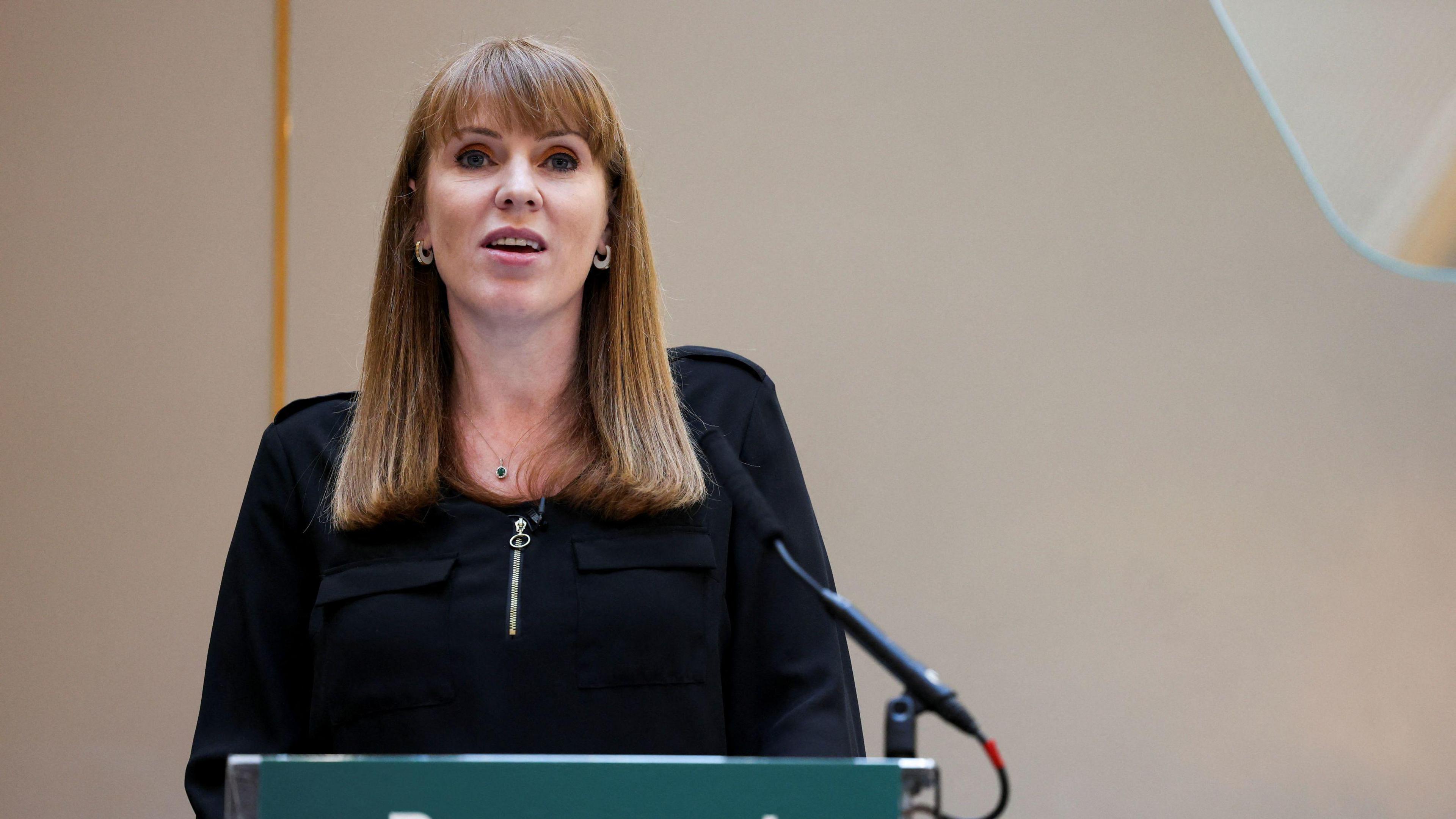 Angela Rayner speaking at an event in Leeds. She is standing behind a dark green lectern into a microphone and is looking out at the audience. She has mid-length ginger hair with a thick fringe and is wearing pink eyeshadow. She also wears hoop earrings and a long-sleeve black top or dress with a prominent zip on the front. 