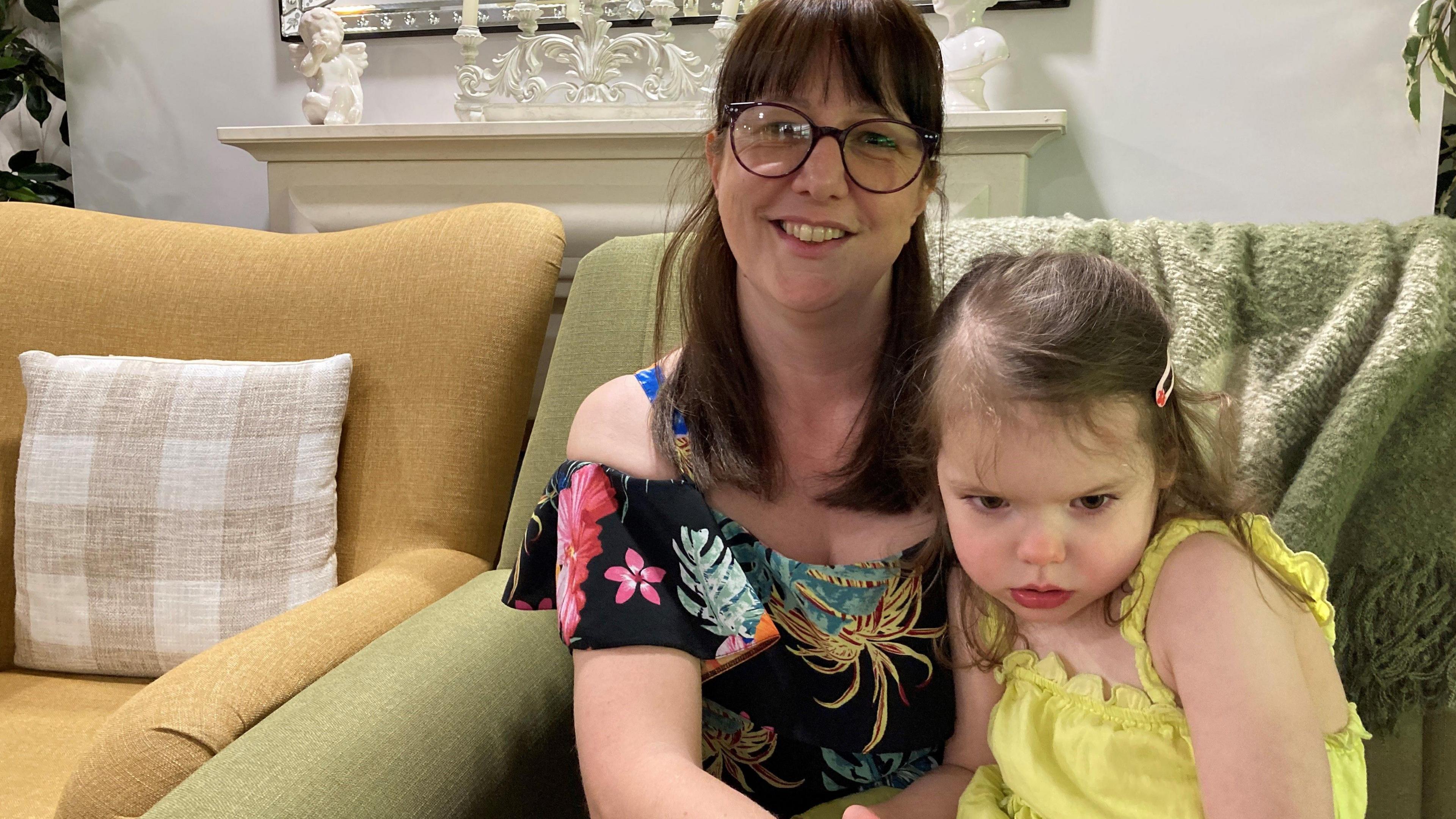A smiling lady in brightly coloured top and glasses holds a toddler wearing a lemon-yellow outfit. They sit on a sofa with a light green covering in front of a cream-coloured fireplace.