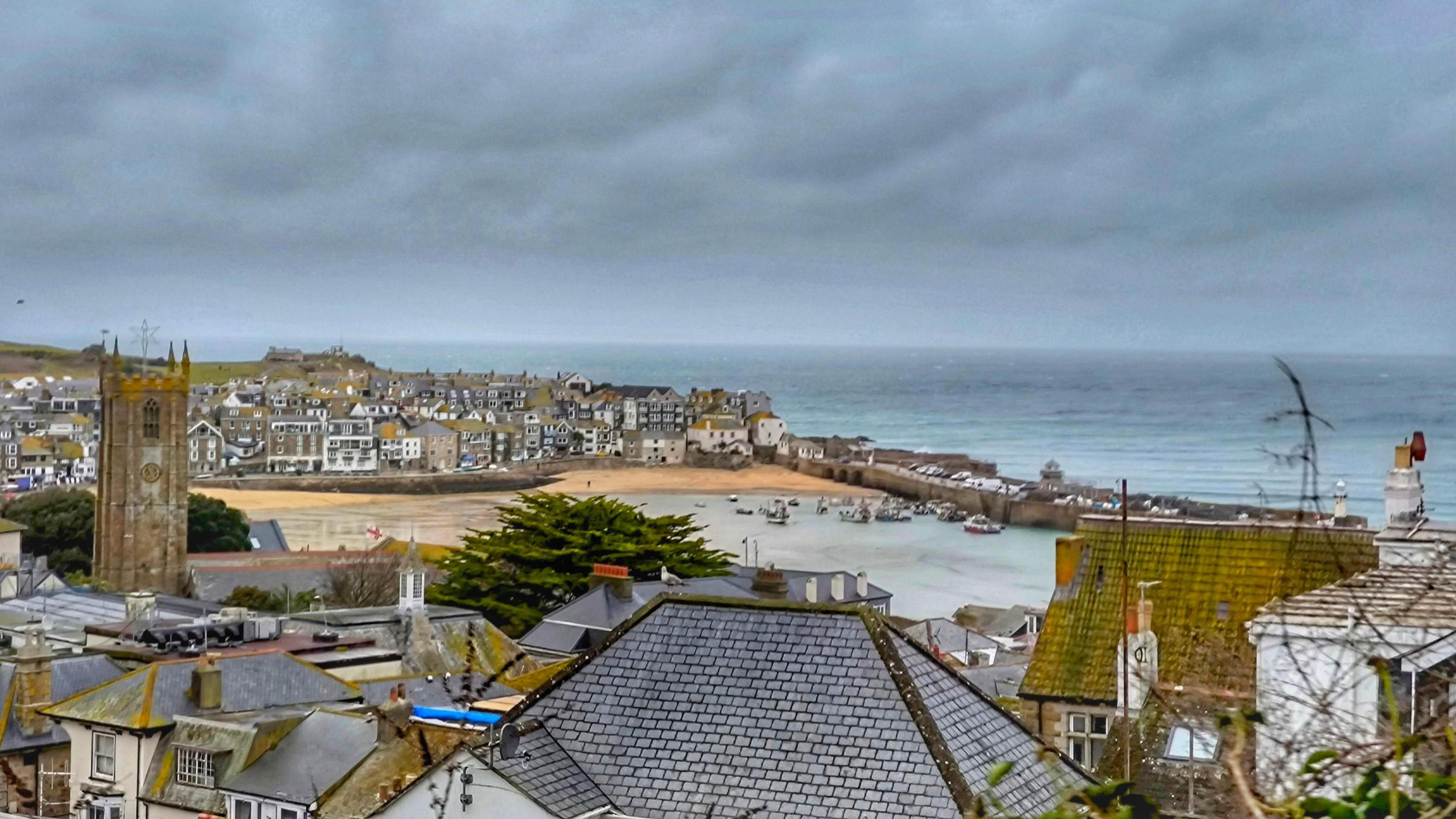 St Ives in Cornwall on an overcast day. There is a large band of grey cloud in the sky. The town's harbour is in the distance and there are several boats on the water.