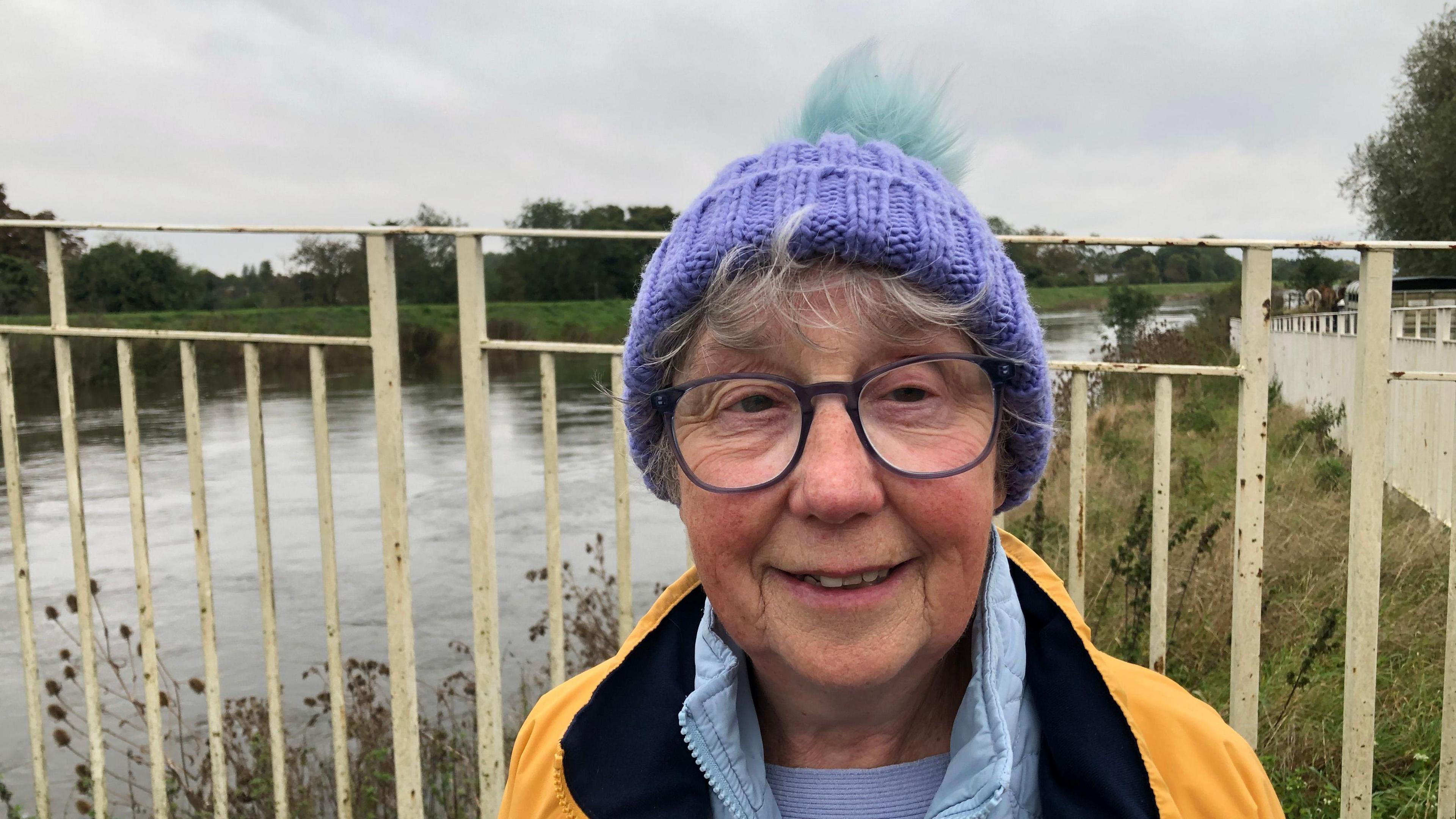 Pat Leavesley has a wisp of white hair forming a fringe from under a violet woollen bobble hat. She is wearing large framed glasses, a lilac under shirt, light blue fleece and a yellow overcoat with black lining. Behind her is the swollen River Great Ouse.