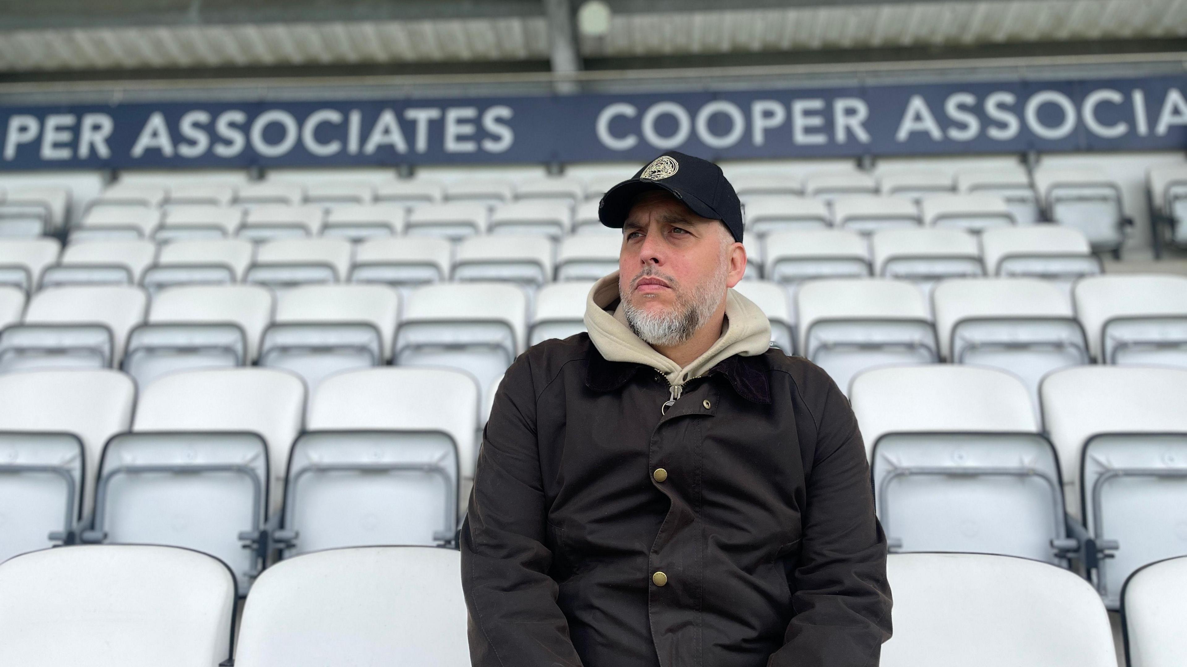 Daniel James sits on tiered seating at a cricket ground. He is wearing a dark jacket, beige hoodie and a black cap.