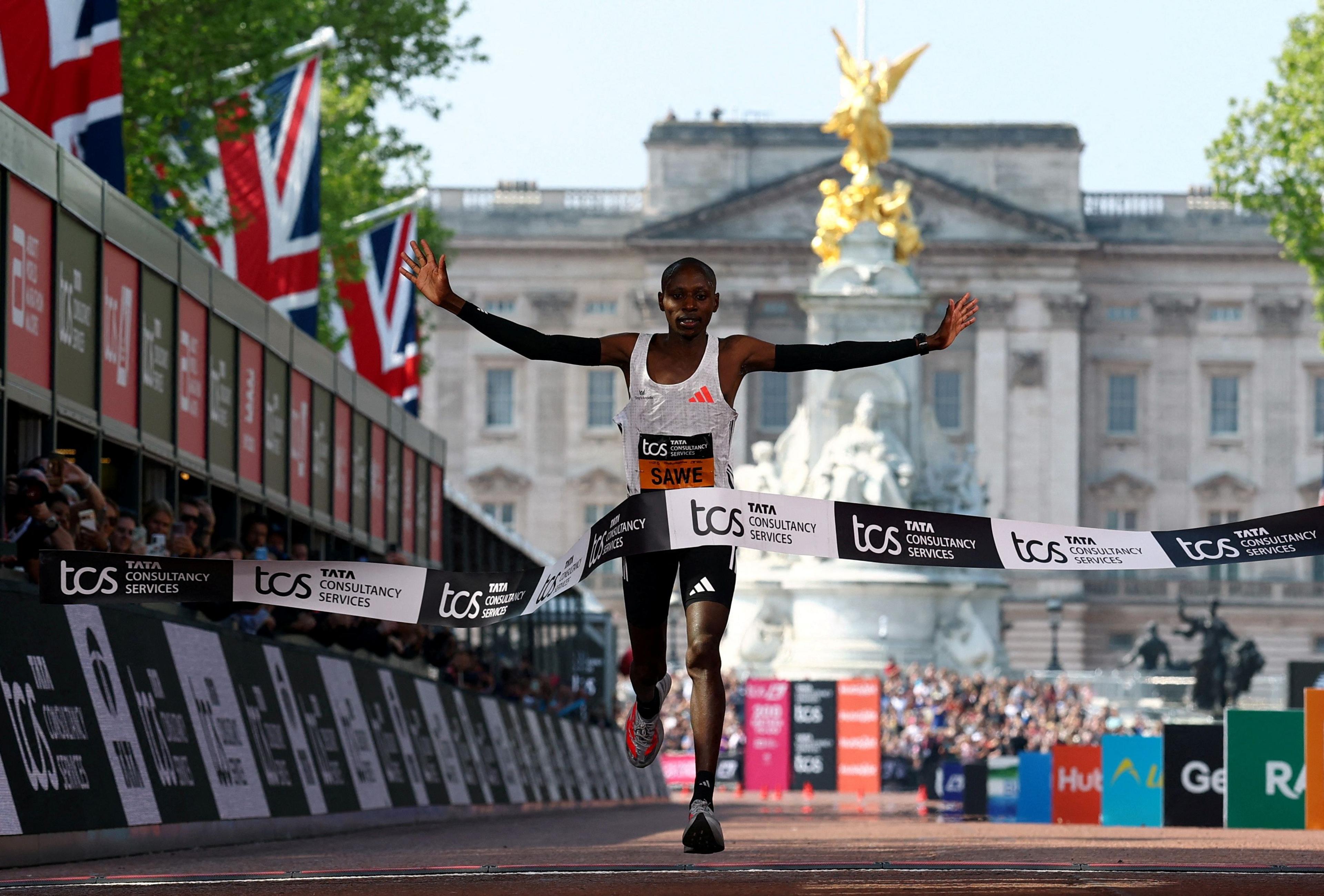 Kenya's Sabastian Sawe crosses the finish line to win the men's elite race. The ribbon falls as he runs through it, and Buckingham Palace is visible in the background.