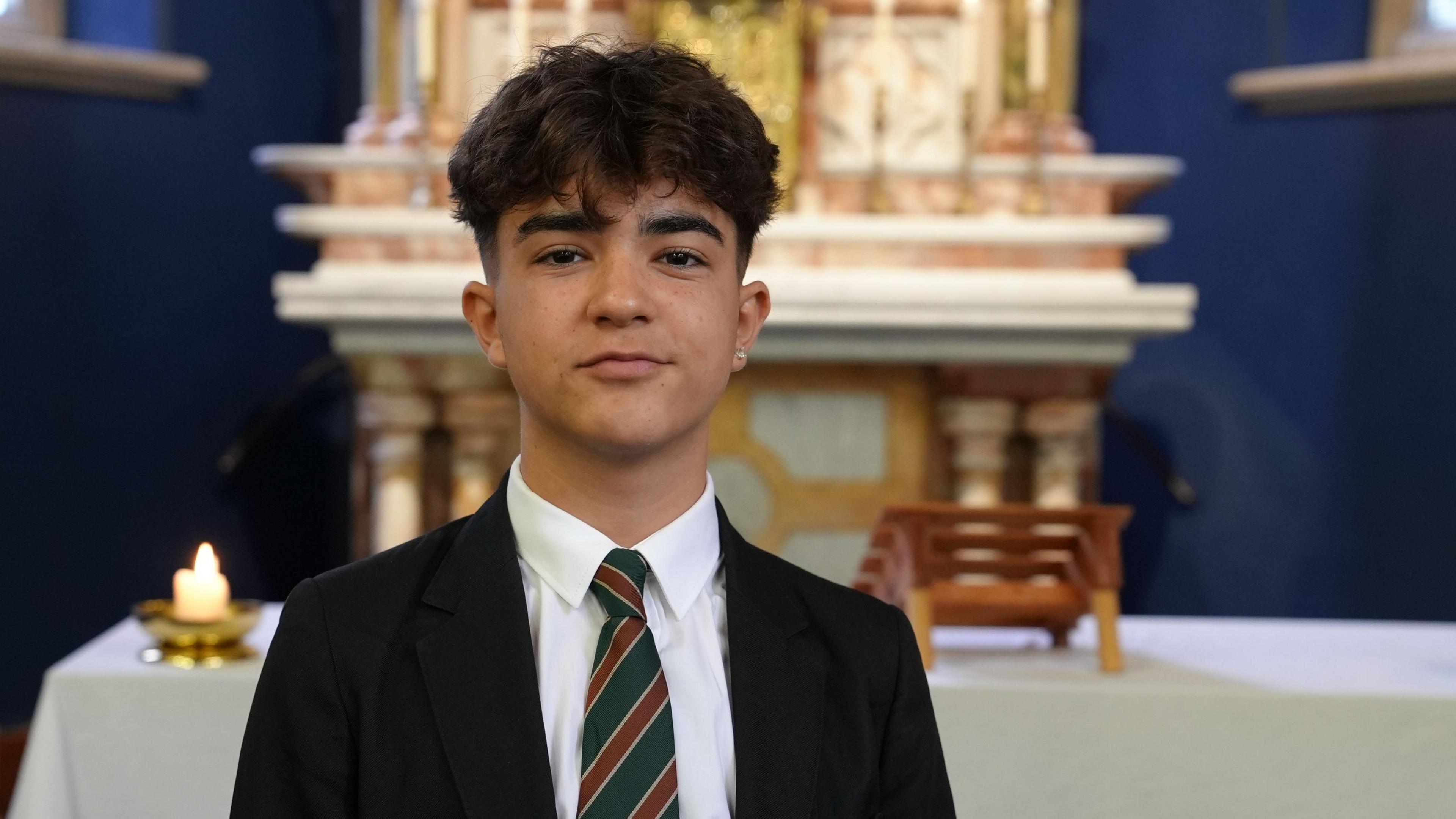 Luis is a teenage boy dressed in a black school blazer, diagonally striped tie and white shirt. He is inside a church with an alter behind him.
