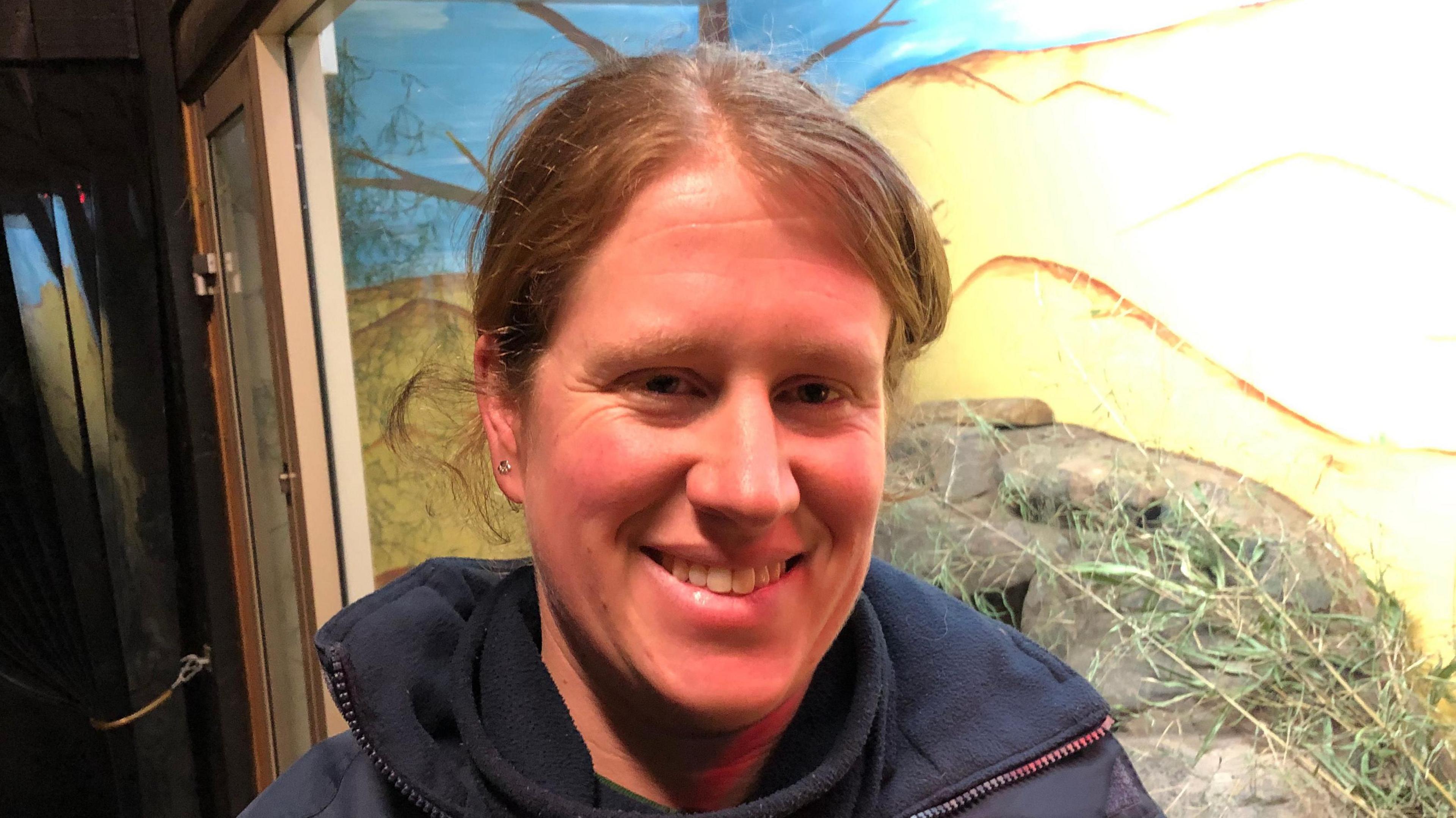 Zoe Nunn with light brown hair smiles at the camera. She is wearing a blue coat with a blue top underneath. She is standing in front of an animal enclosure which has a glass window front.