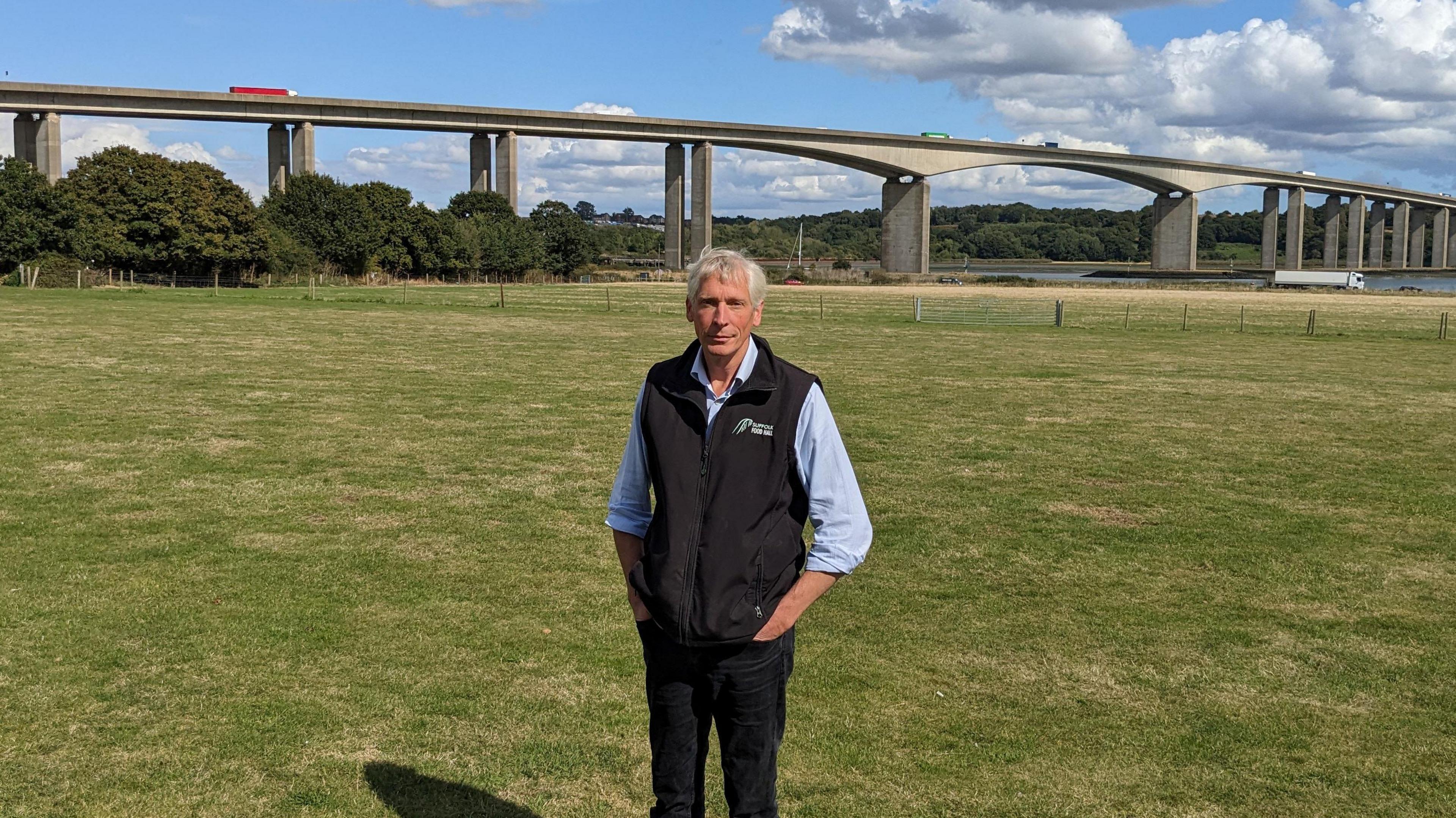 Oliver Paul, standing on grass, with the River Orwell and the Orwell Bridge behind him