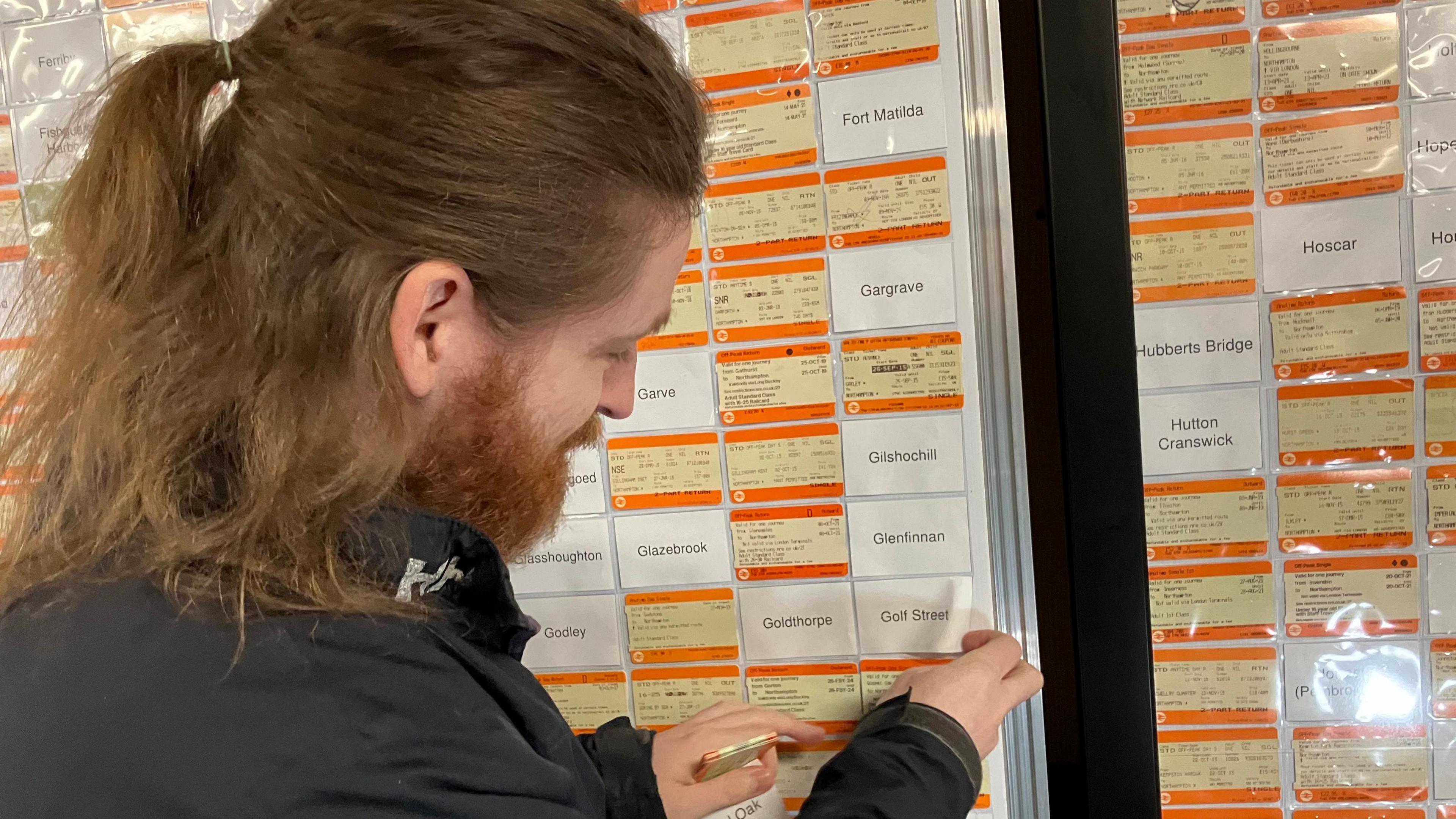 Elliott Badger who has should-length brown hair pulled back from his forehead and a beard. He is wearing a black jacket and standing facing of a notice board which is packed with railway tickets