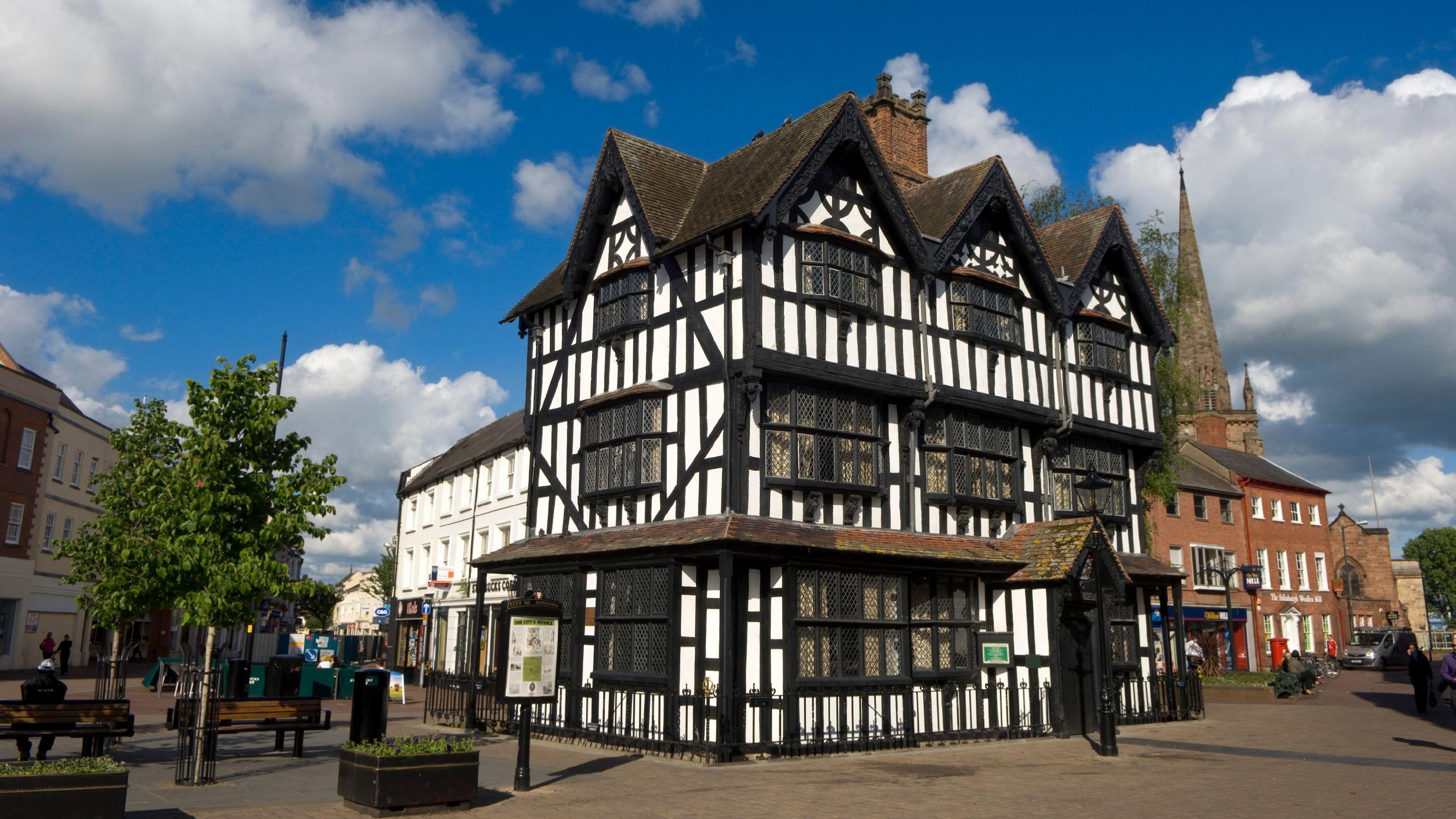 A three-storey tudor building, surrounded by shops. The sky is blue with clouds and there is a church steeple in the right-hand corner of the image.  