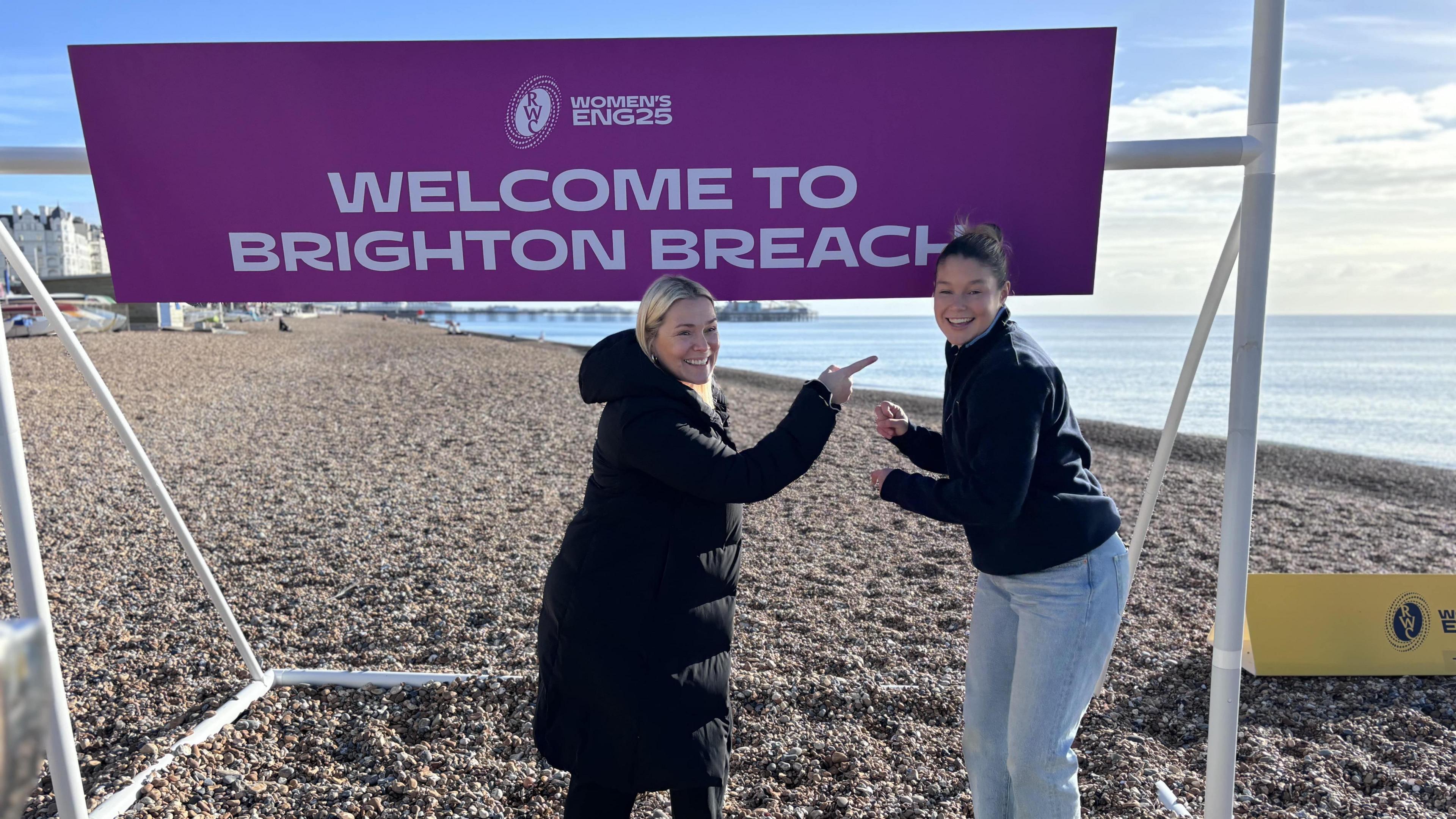 Jess Breach (right, a woman with light blue jeans, a dark blue jumper and her hair in a bun, smiling looking into the camera) and Rachael Burford (left, a woman with blonde hair and a long black coat pointing a Jess) standing in front of a purple sign with white writing on saying Welcome To Brighton Breach on Brighton beach