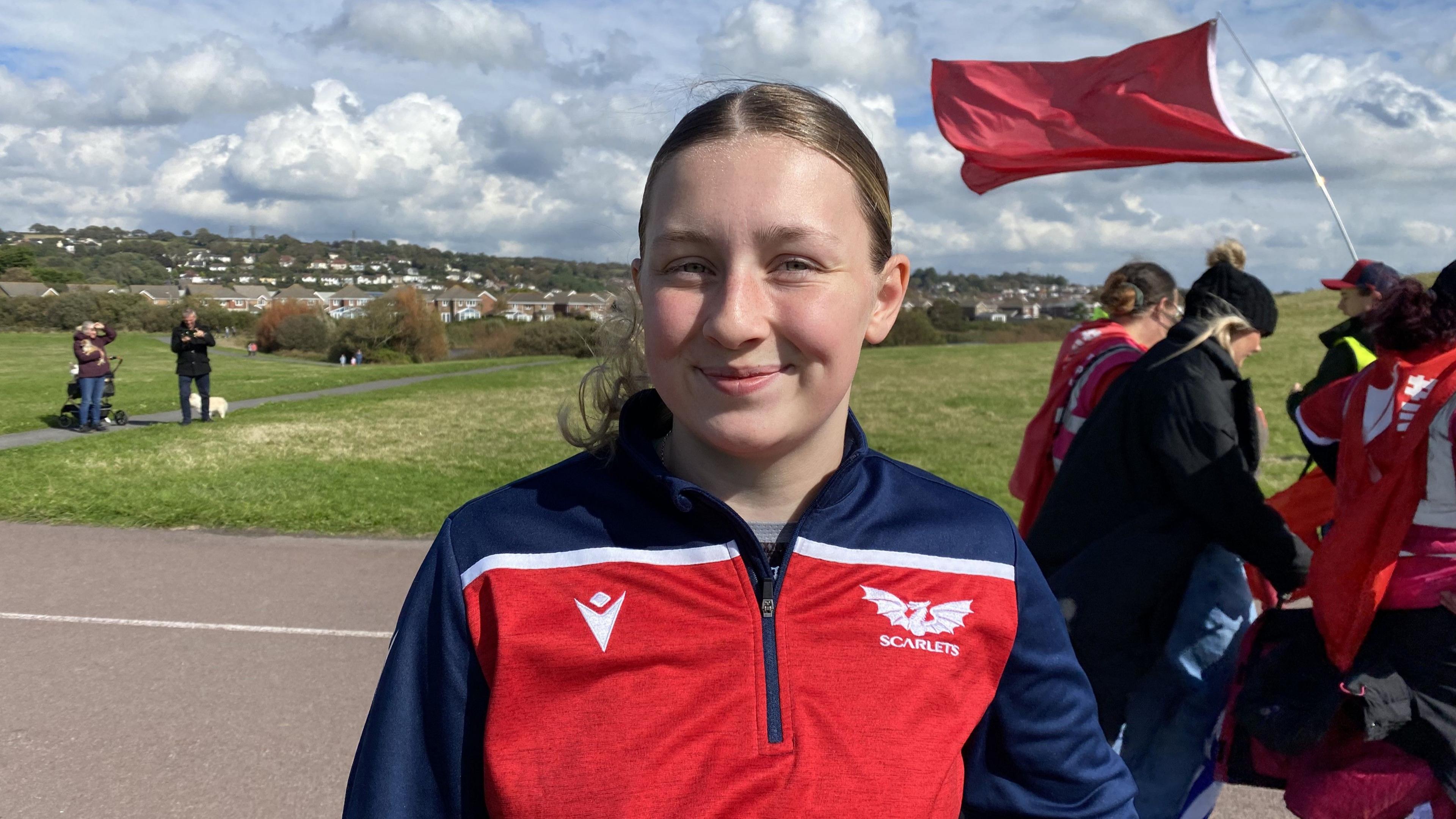 Mae gwallt cara wedi clymu yn'ôl ac mae'n gwenu i'r camera. Mae'n gwisgo crys coch, gwyn a glas tywyll gyda bathodyn y Scarlets. 