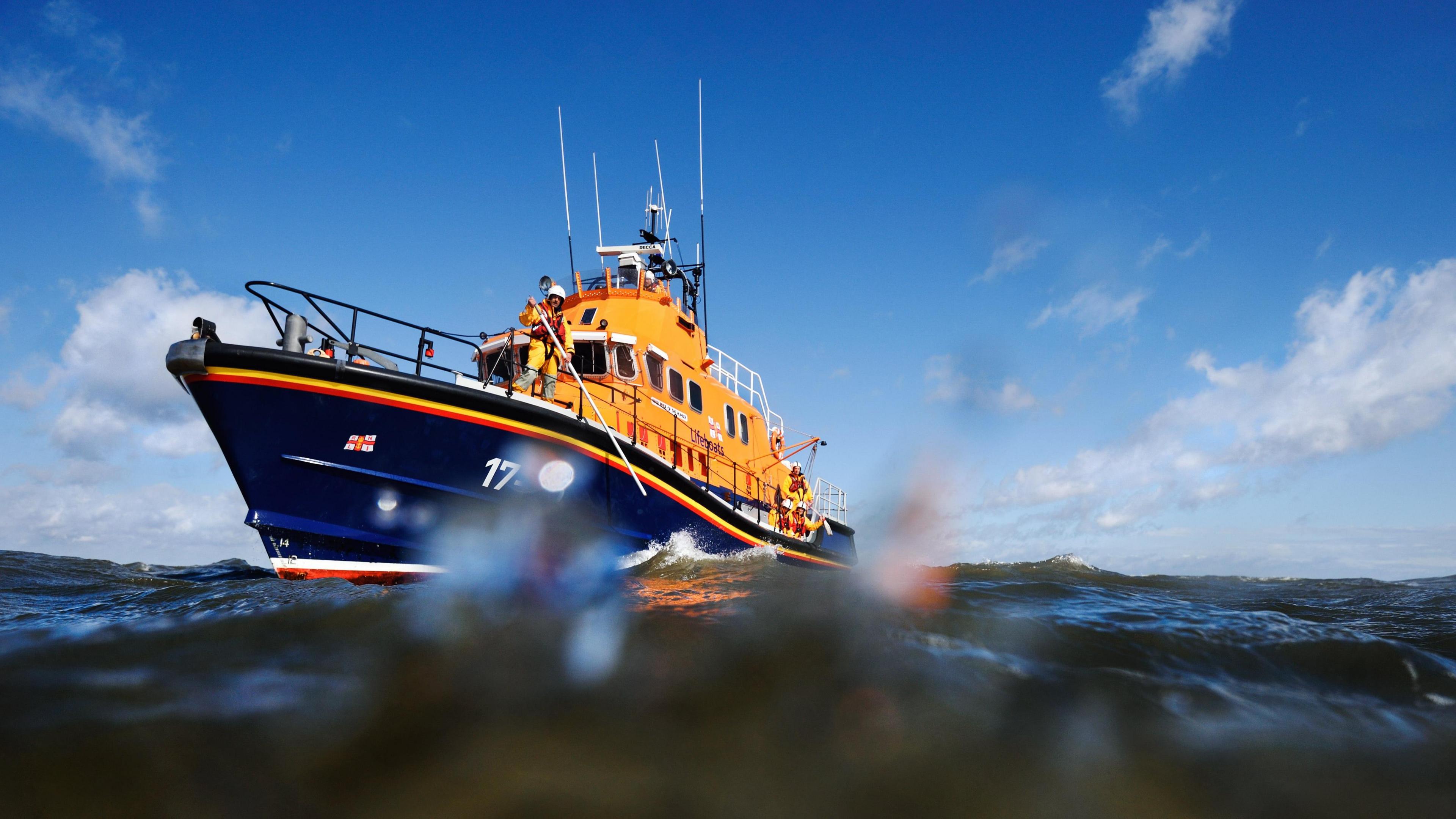 An orange and blue lifeboat, with a member of crew using some rescue equipment. The photo is taken from within the waves. There is a blue sky in the background.