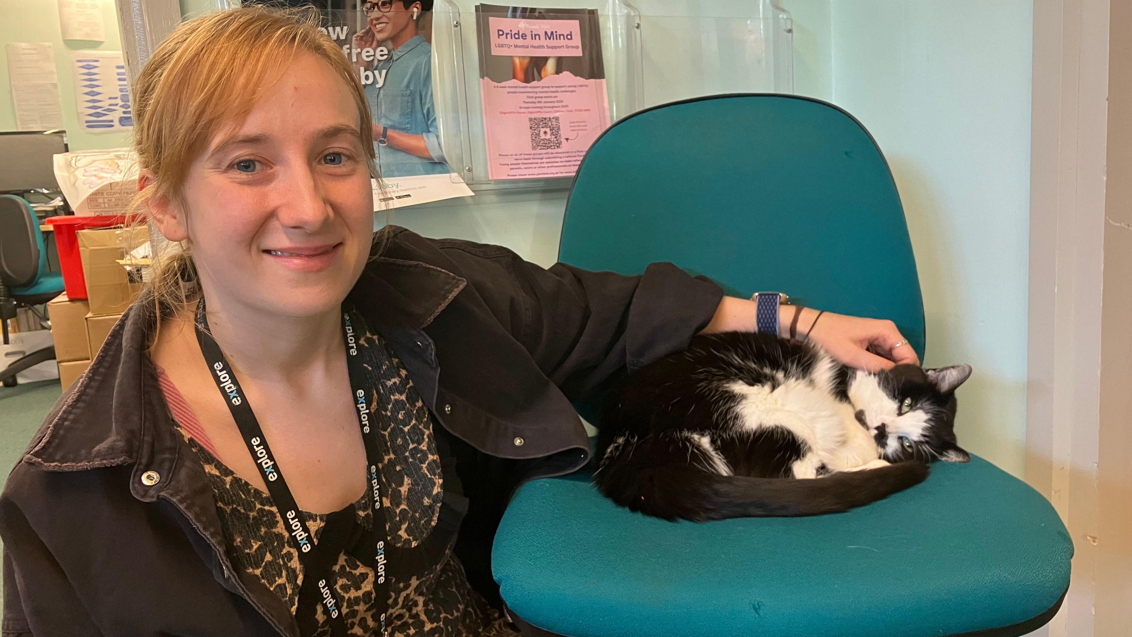 Katie, reading and learning advisor at the library is pictured with Betty the cat curled up on an office chair. She wears a leopard print dress and a black shirt. Betty the cat is black and white.