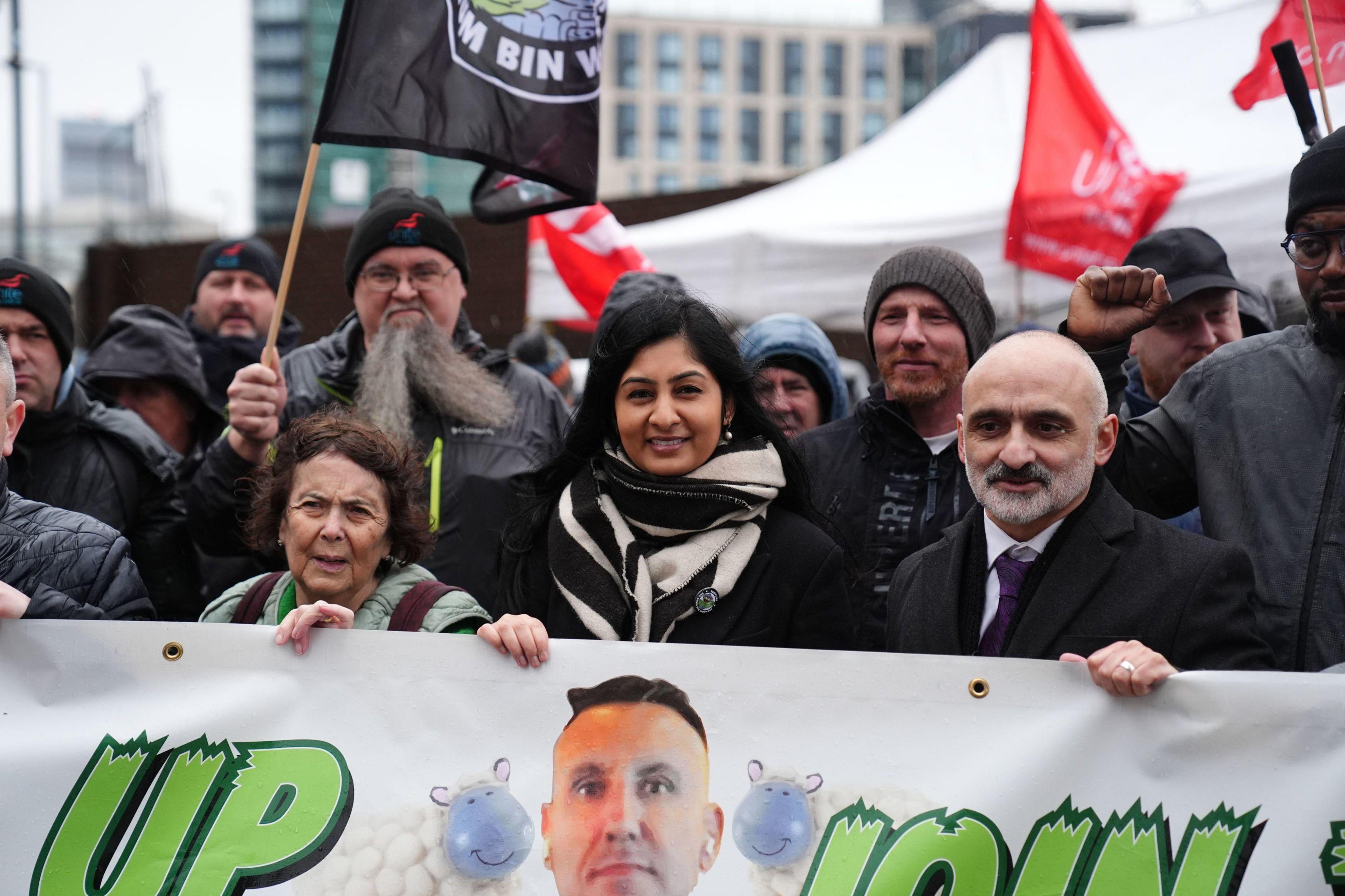 A smiling Zarah Sultana stands behind a banner. She is wearing a black and white scarf. To her left is Unite's national officer Onay Kasab. He has a grey beard and is wearing a white shirt, purple tie and black coat.