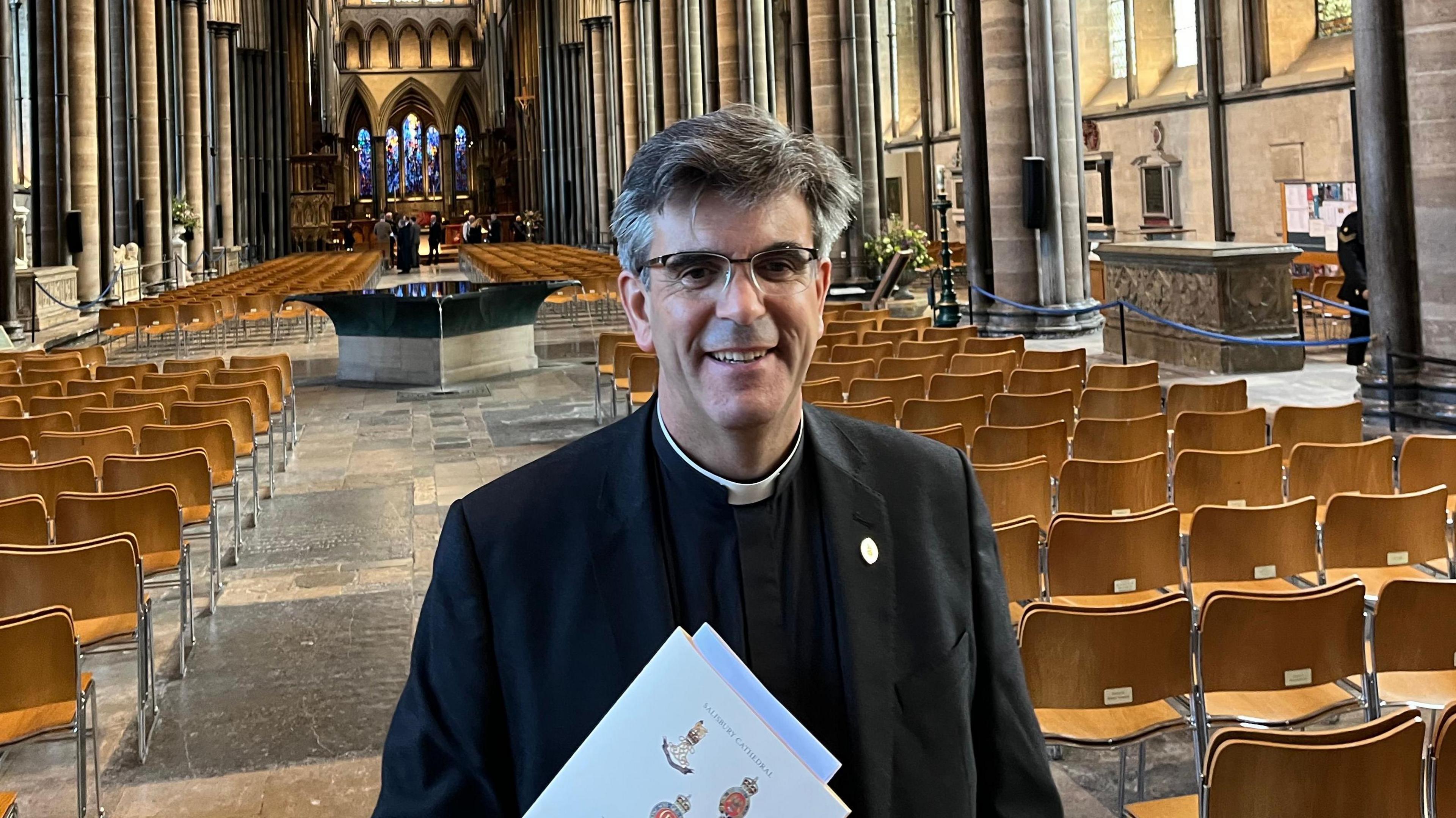 Nick Papadopulos, Dean of Salisbury Cathedral stood by various brown seats with the church facade behind him.
