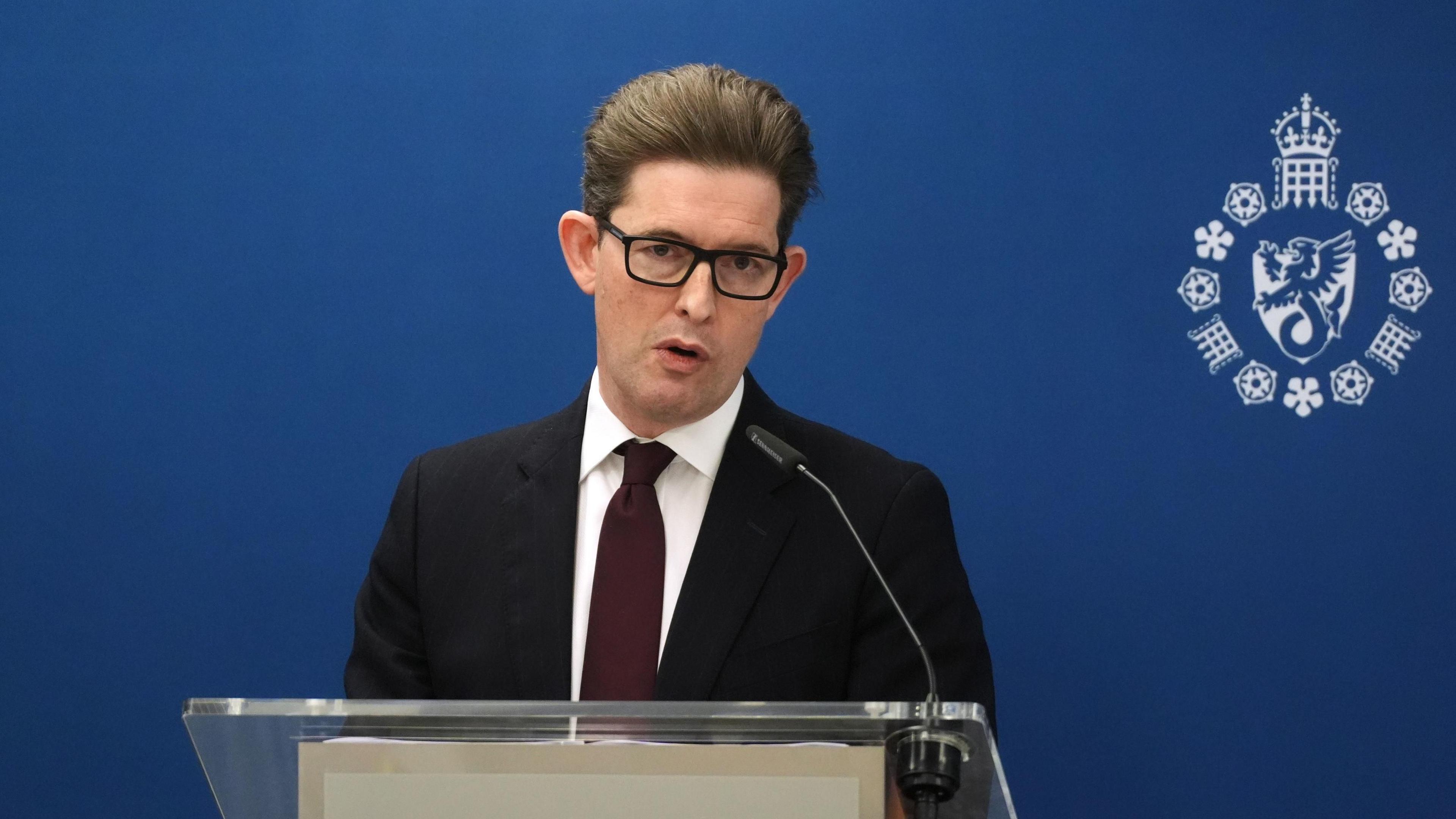 Sir Ken McCallum, delivers his speech. He has short brown hair and wears black-rimmed glasses. He wears a black suit jacket, white shirt and dark red tie. He is speaking into a small microphone on a glass lectern, in front of a blue wall.
