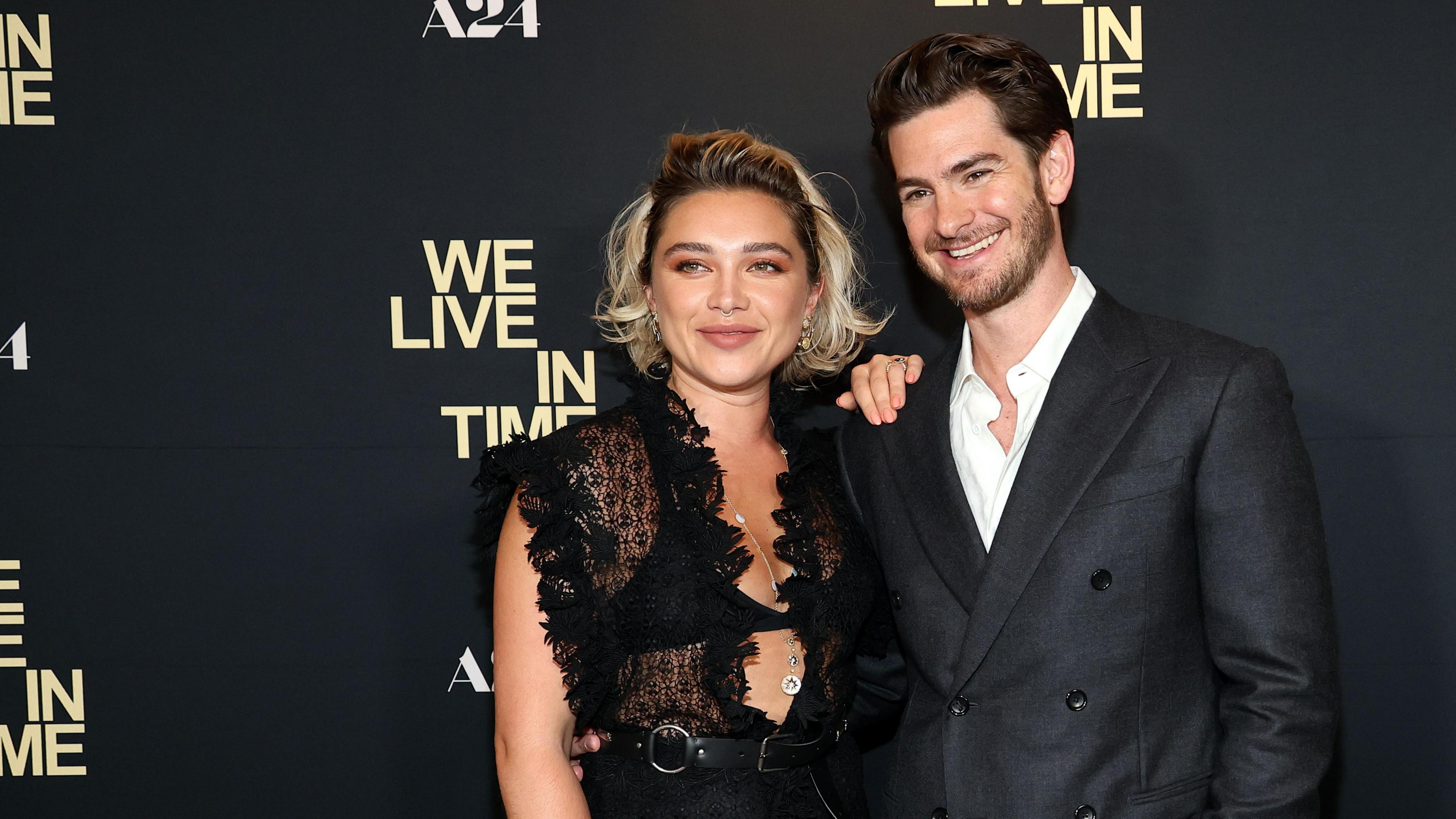 Florence Pugh in a black lace outfit with Andrew Garfield in a black suit standing together and smiling against a background that says 'We Live In Time'. 