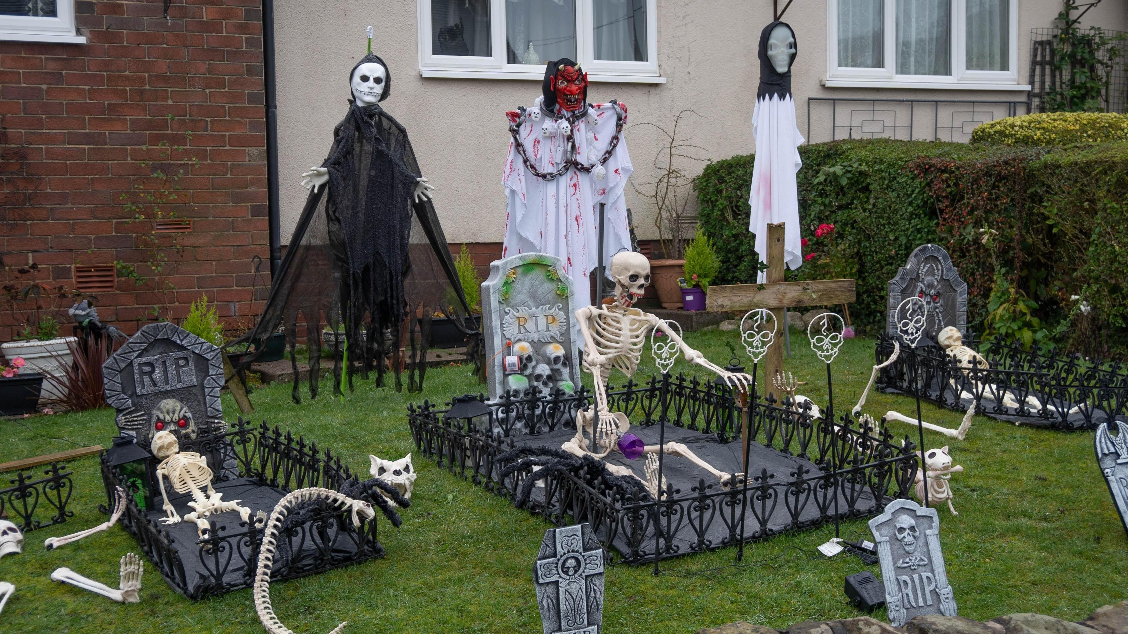 A Halloween display in the front garden of a partly rendered house. It includes a number of fake gravestones, some with decorative fences around them, skeletons in human and animal forms, and three ghostly characters against the back wall. 